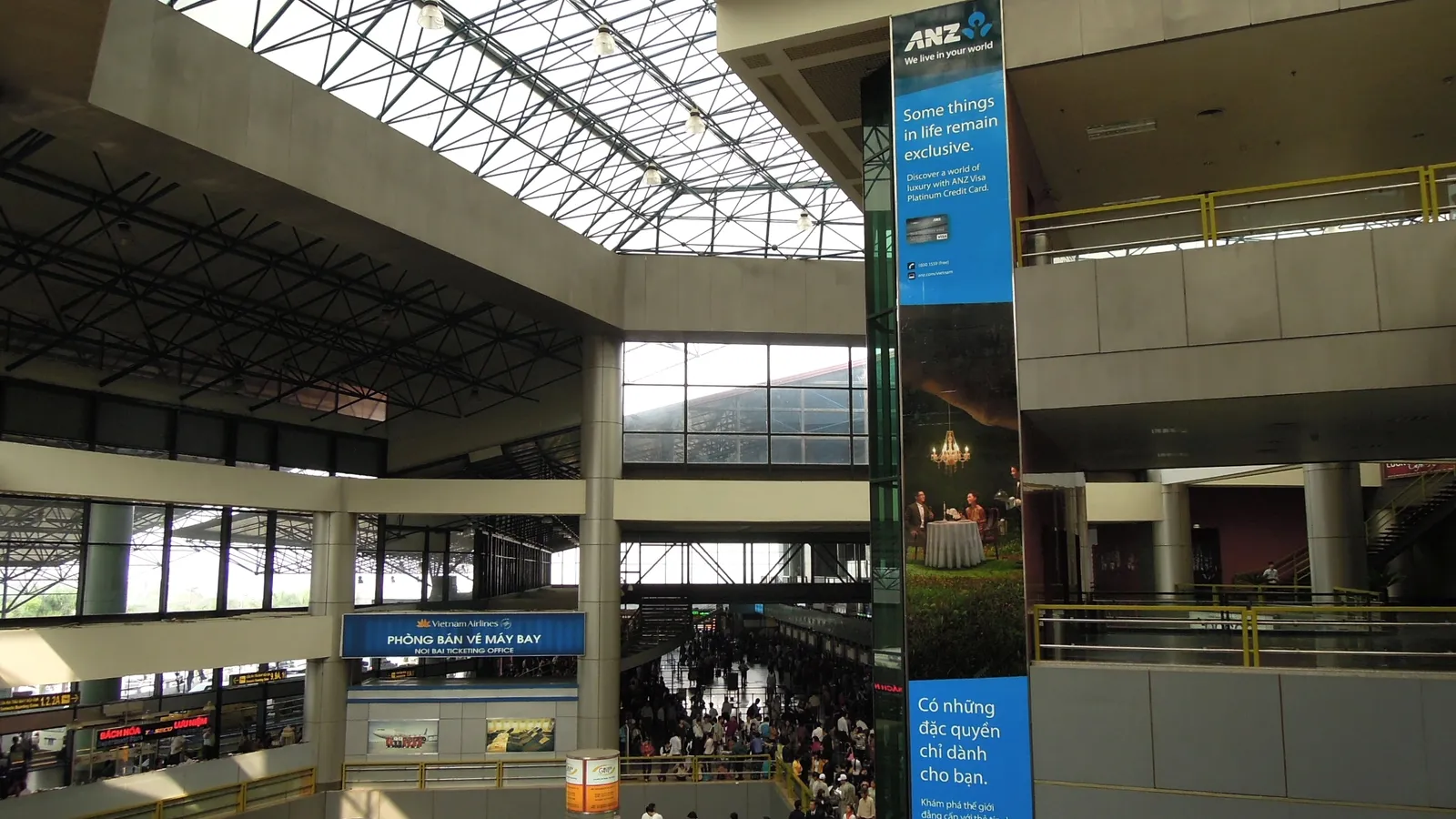 The public concourse inside a terminal at Hanoi's Noi Bai airport, a tall glass-roofed hall with escalators, a ticket counter and passengers queueing on the floor below