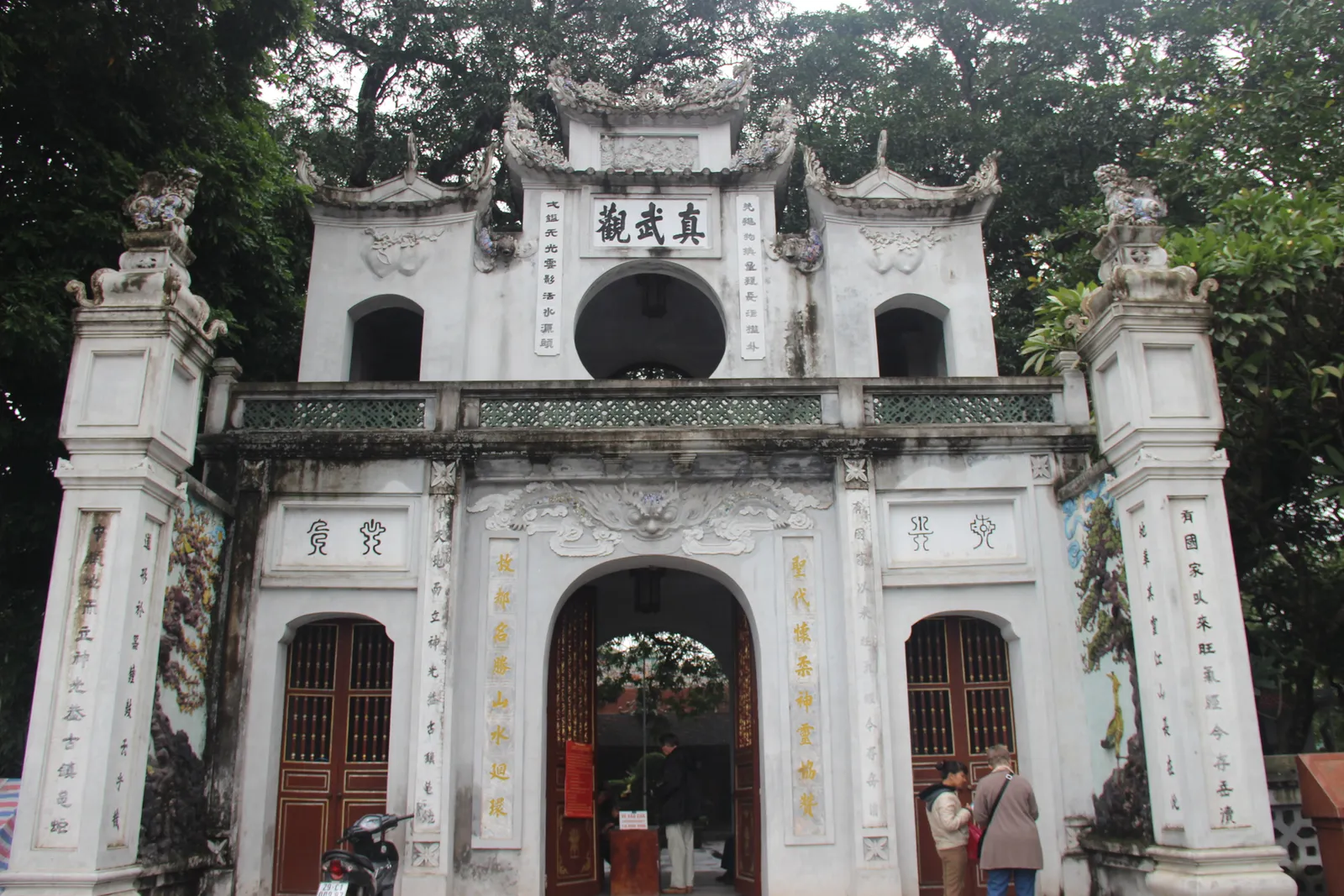 The whitewashed triple gateway of Quan Thanh Temple in Hanoi, carved Chinese characters across its upper panel and stone dragons on the roofline, with visitors passing through the central arch