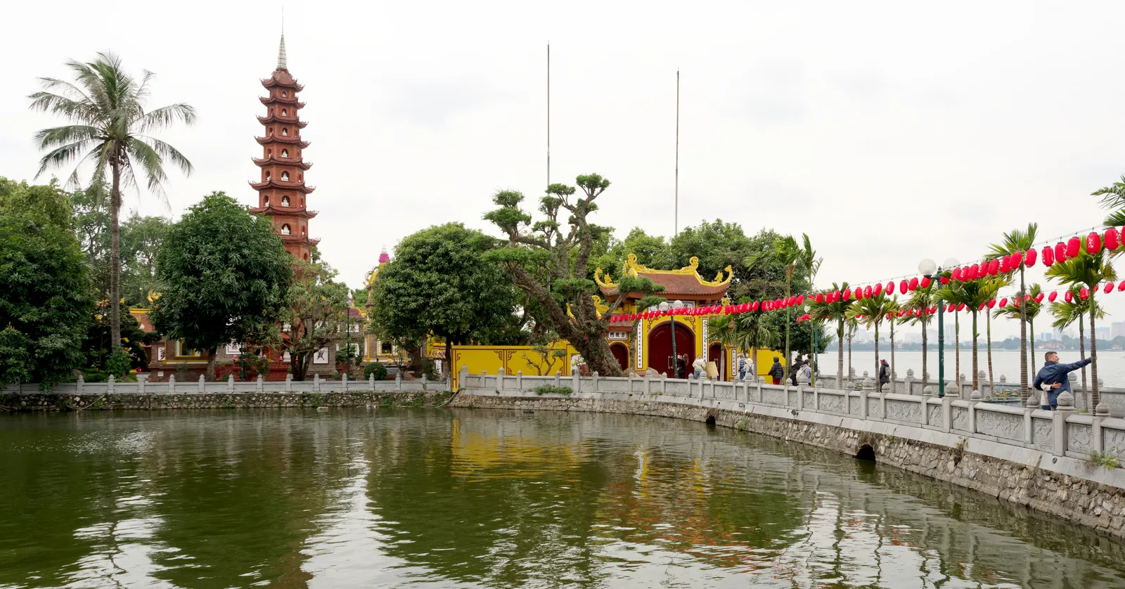 Tran Quoc Pagoda on its islet in West Lake, Hanoi, an eleven-tier red brick stupa rising above yellow temple walls, with a railed causeway and strings of red lanterns along the water's edge