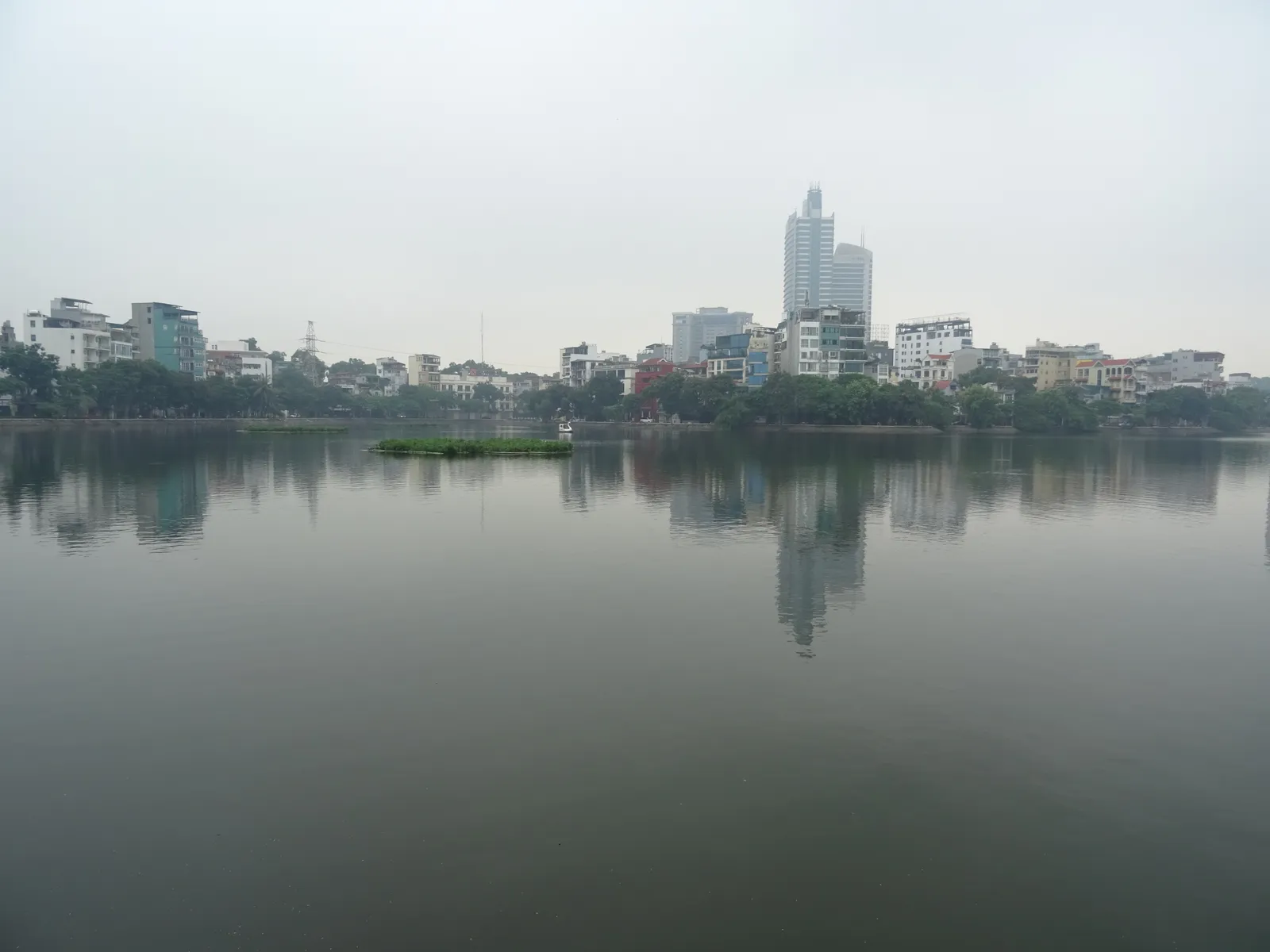 Truc Bach Lake in Hanoi on an overcast day, flat grey water reflecting the low-rise houses and a single tall tower along the far shore