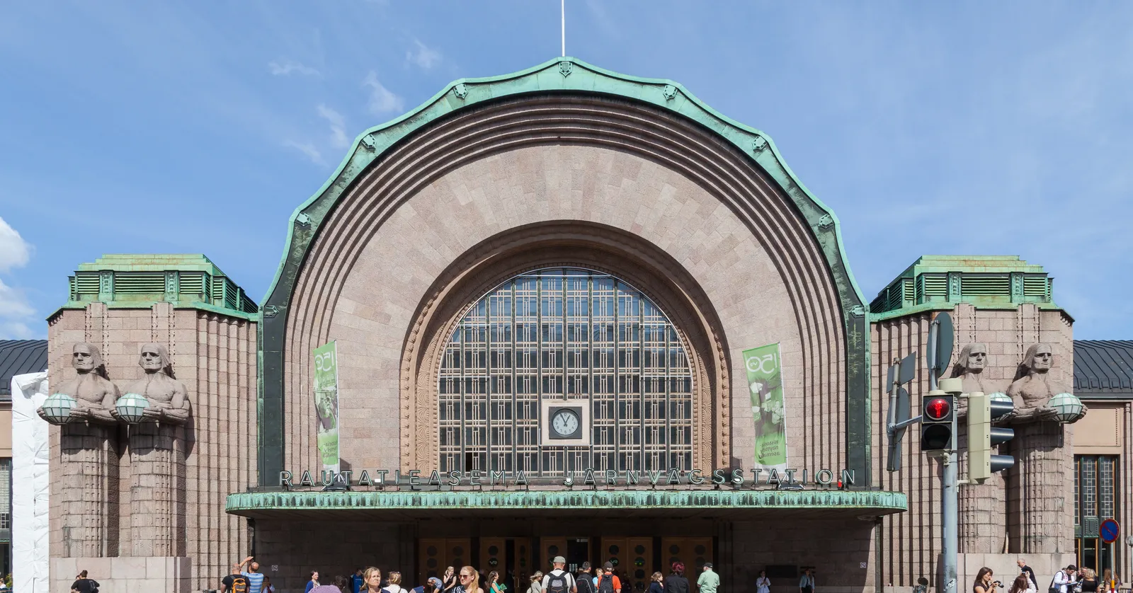 The main entrance of Helsinki Central Station, a huge arched window set in a facade of pink granite, with two pairs of giant seated stone figures holding round lamps standing either side of the doors