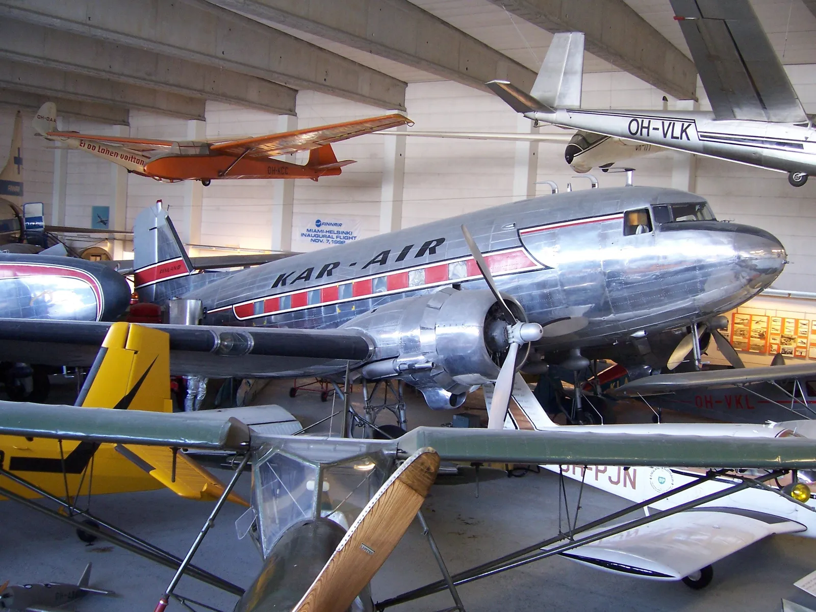 Inside a hangar at the Finnish Aviation Museum in Vantaa, a polished bare-metal DC-3 airliner in red and silver livery parked among light aircraft and gliders, with more planes hung from the roof beams