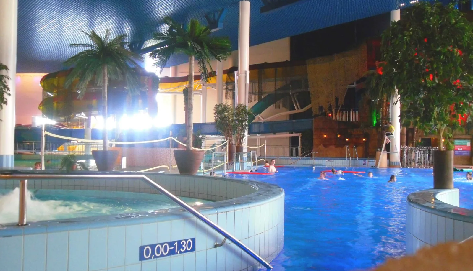 The indoor waterpark at Flamingo in Vantaa, a large blue pool under a dark vaulted roof with artificial palm trees around it, a raised circular hot pool in the foreground and a water slide curving down at the back
