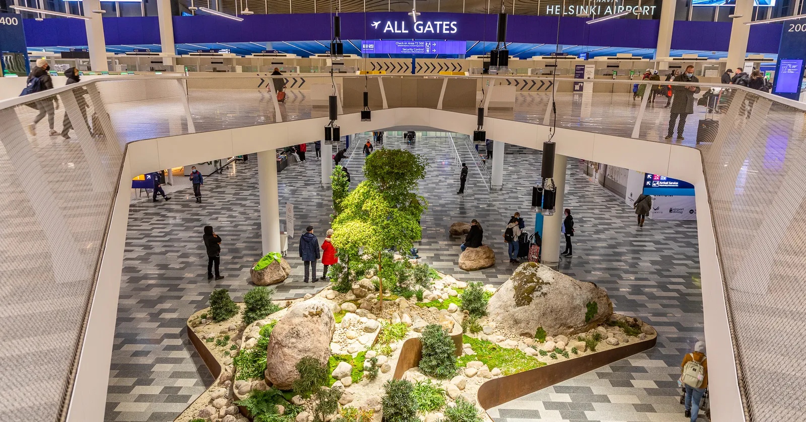 The departures hall at Helsinki Airport seen from a balcony, a wide pale floor with an island of boulders, moss and small pine trees planted in the middle of it, and an overhead sign reading All Gates
