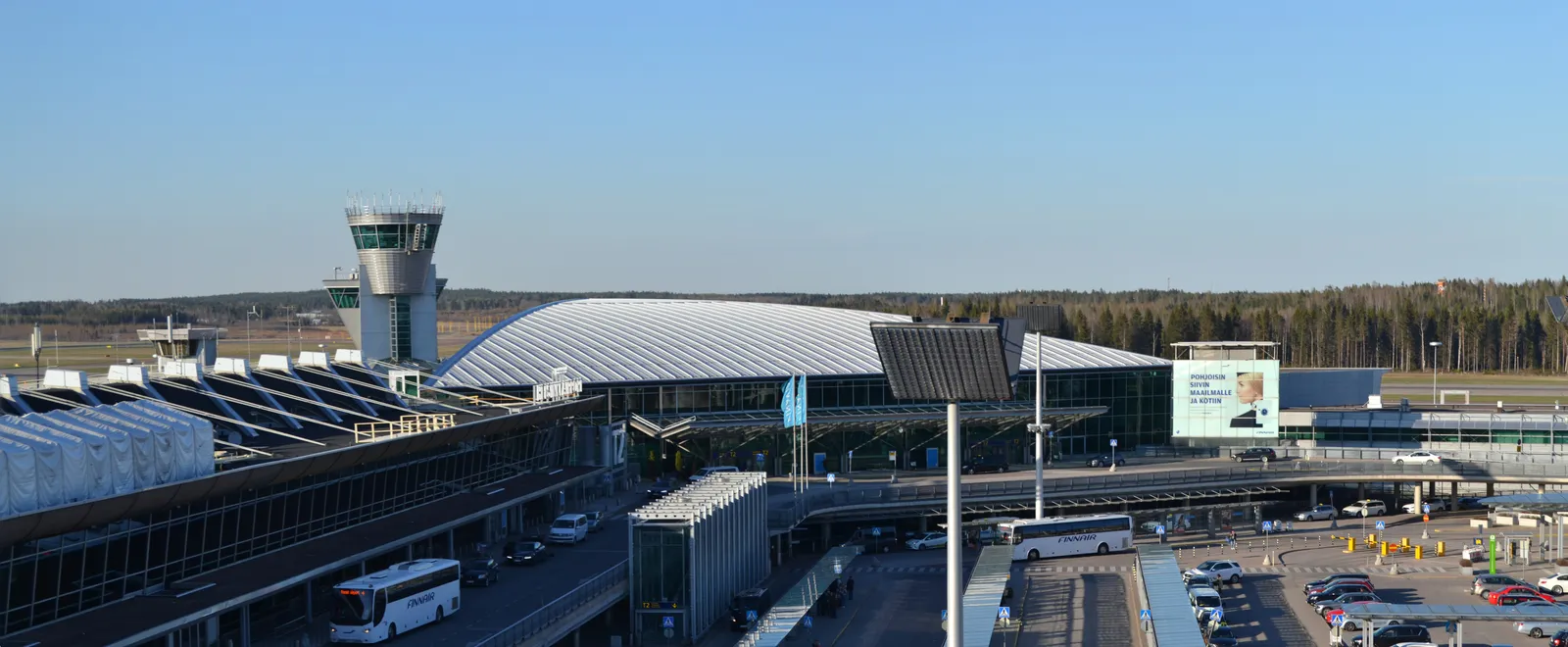 Helsinki Airport seen from a raised terrace, the long curved white roof of Terminal 2 across the road with the control tower behind it, buses and parked cars in front, and forest running along the horizon