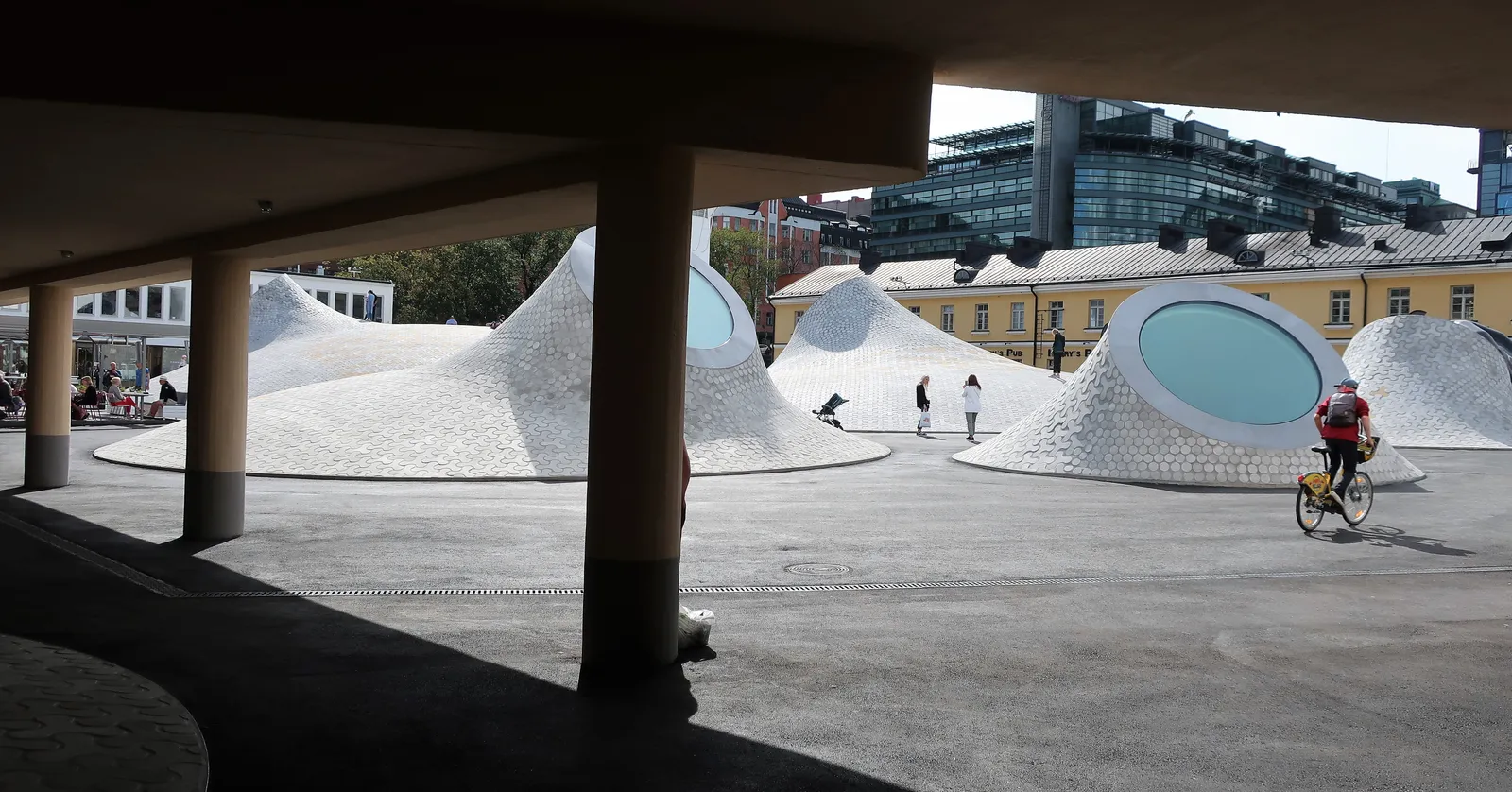 Lasipalatsi square in Helsinki seen from under a colonnade, with several white concrete mounds rising out of the paving, each capped by a round tilted skylight, and people walking up and over them