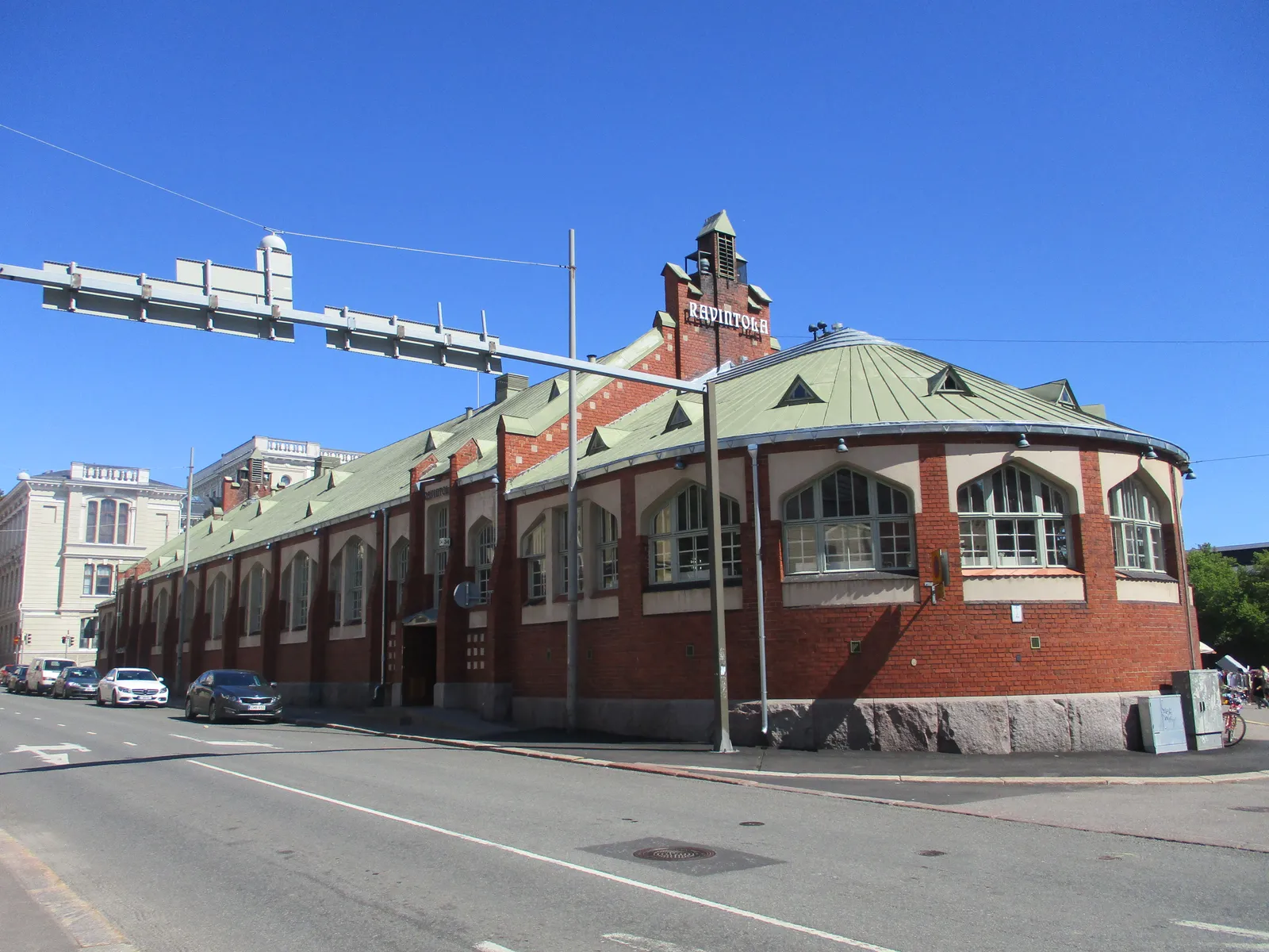 The Hietalahti market hall in Helsinki from the street corner, a long red brick building on a granite base with a green metal roof, arched windows down its side and a small tower over the entrance