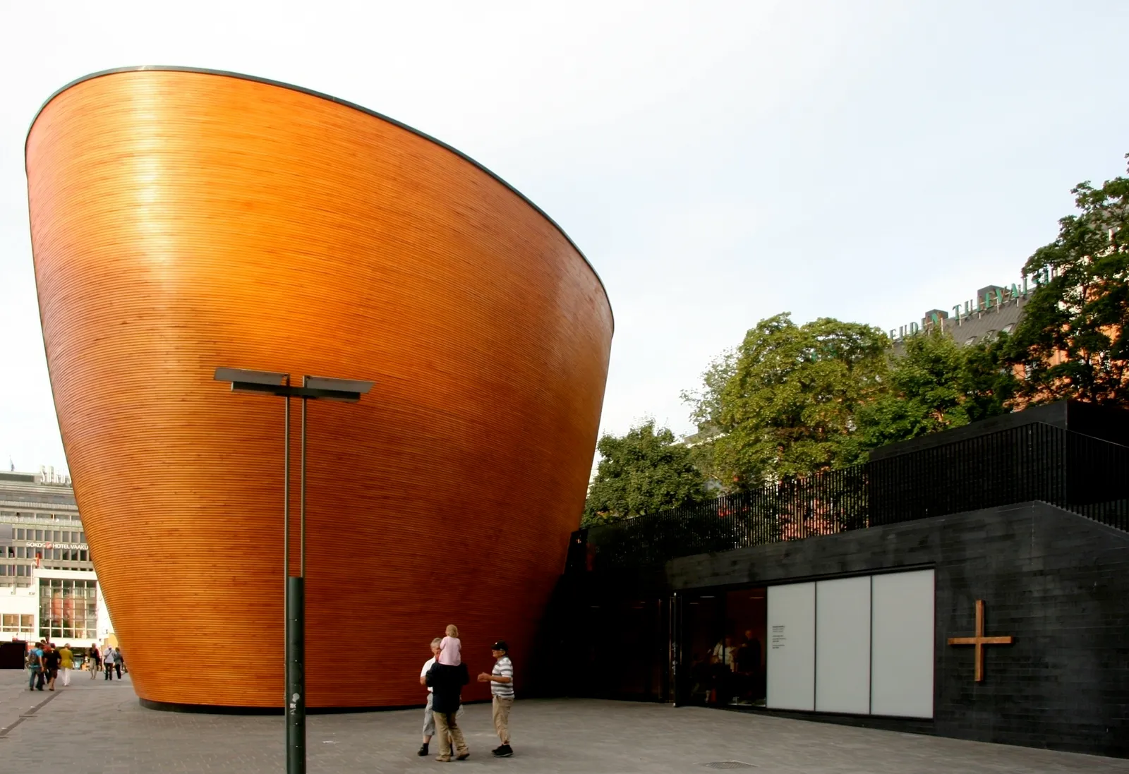 The Kamppi Chapel of Silence in Helsinki, a tall curved bowl-shaped building clad in smooth pale timber staves, standing on an open paved square beside a low dark entrance block with a plain wooden cross on it