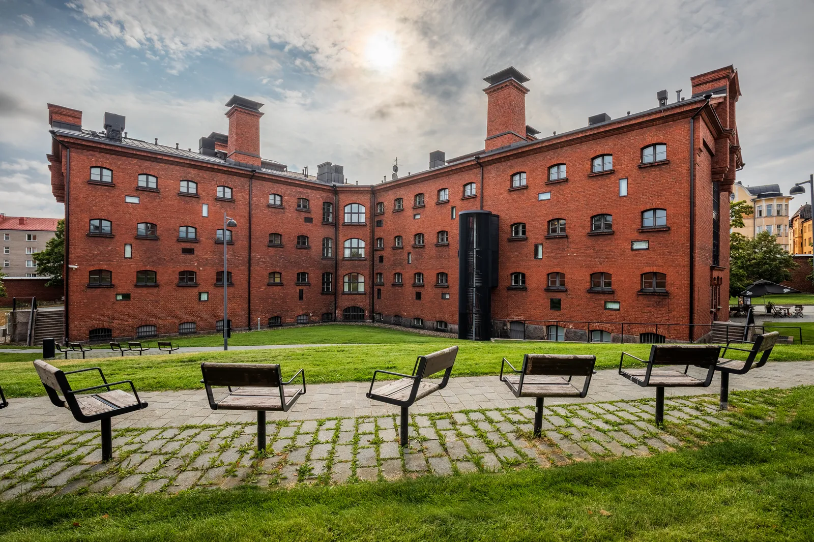 The former Helsinki County Prison on Katajanokka, a long red brick building of three storeys with small regularly spaced windows and tall chimneys, set back behind a lawn with benches on a paved strip in front
