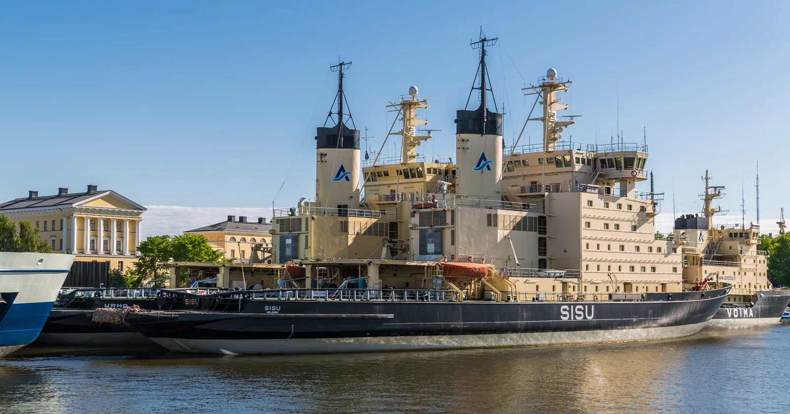 Three Finnish icebreakers moored side by side along the Katajanokka quay in Helsinki, black hulls with the names Sisu, Urho and Voima on the bow, cream superstructures and funnels above them, and a pale colonnaded warehouse on the quayside behind