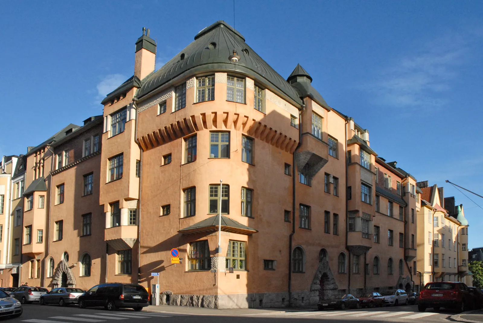 An apartment block on a Katajanokka street corner in Helsinki, apricot-rendered walls under a steep green roof, with a rounded corner bay, a small turret, rows of bracket-like details under the eaves and a granite base at street level