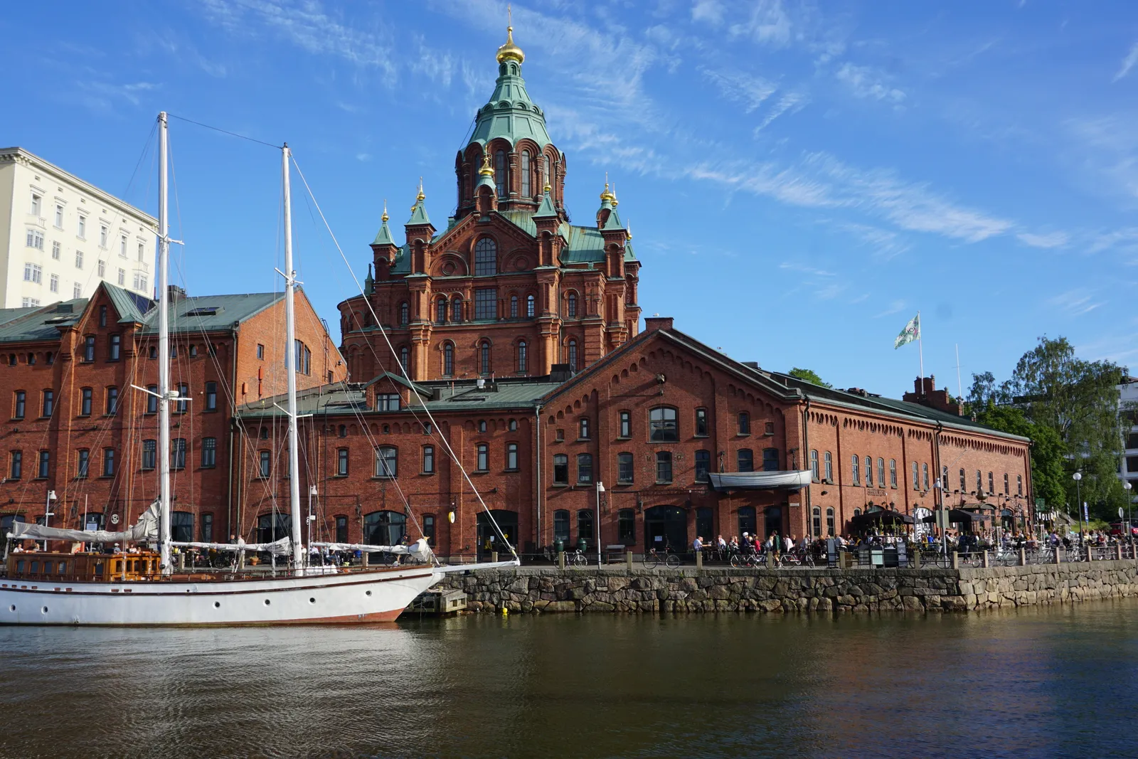 Uspenski Cathedral in Helsinki seen from across the water, red brick walls and green copper domes topped with gold crosses rising behind a row of long red brick warehouses on the Katajanokka quay, with a white wooden schooner moored in front
