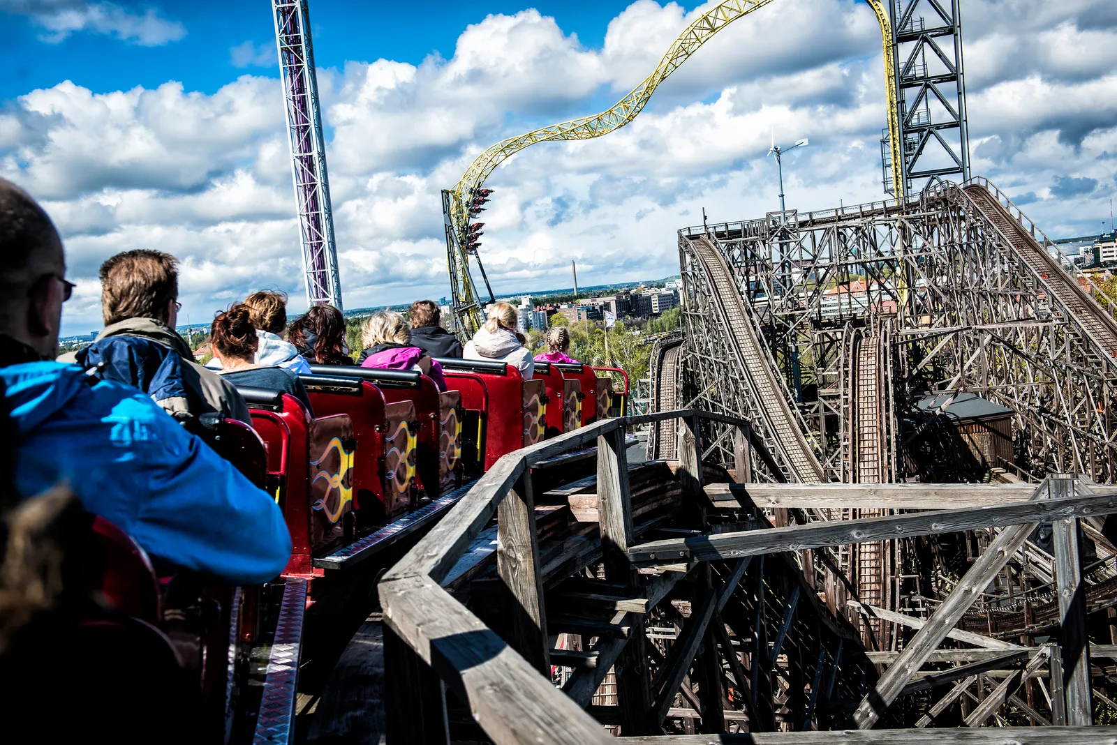The view from a seat on the wooden roller coaster at Linnanmäki in Helsinki, riders in a red train on a timber track curving between weathered wooden trestles, with a yellow steel coaster looping against a blue sky behind