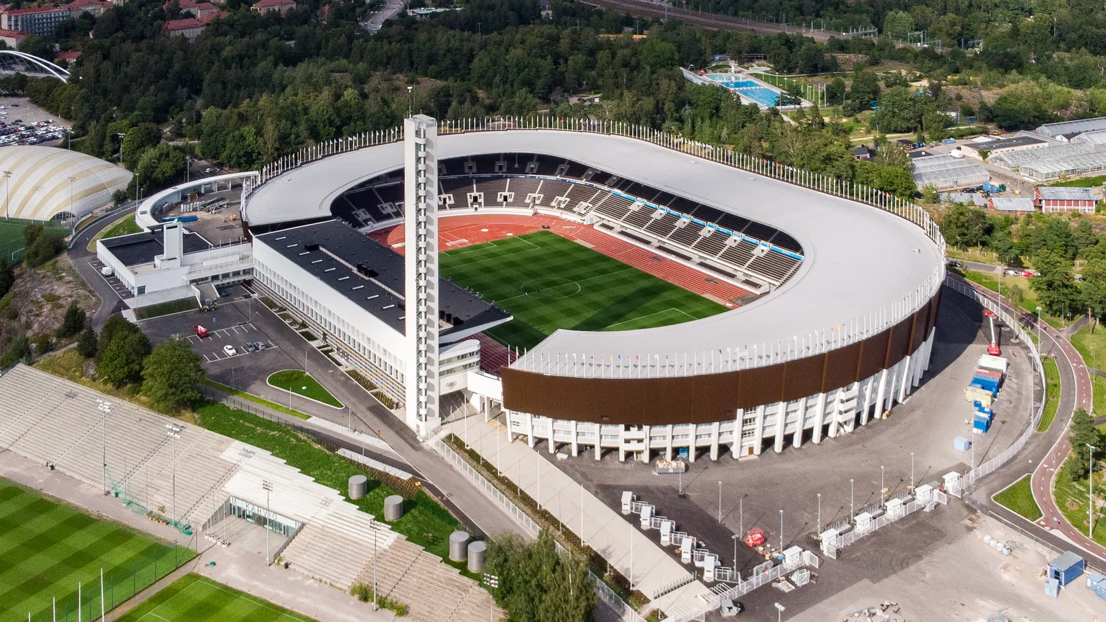 The Helsinki Olympic Stadium from the air, a white oval of curved stands around a green pitch and a red running track, with a slim white tower standing beside the western stand and woodland on all sides