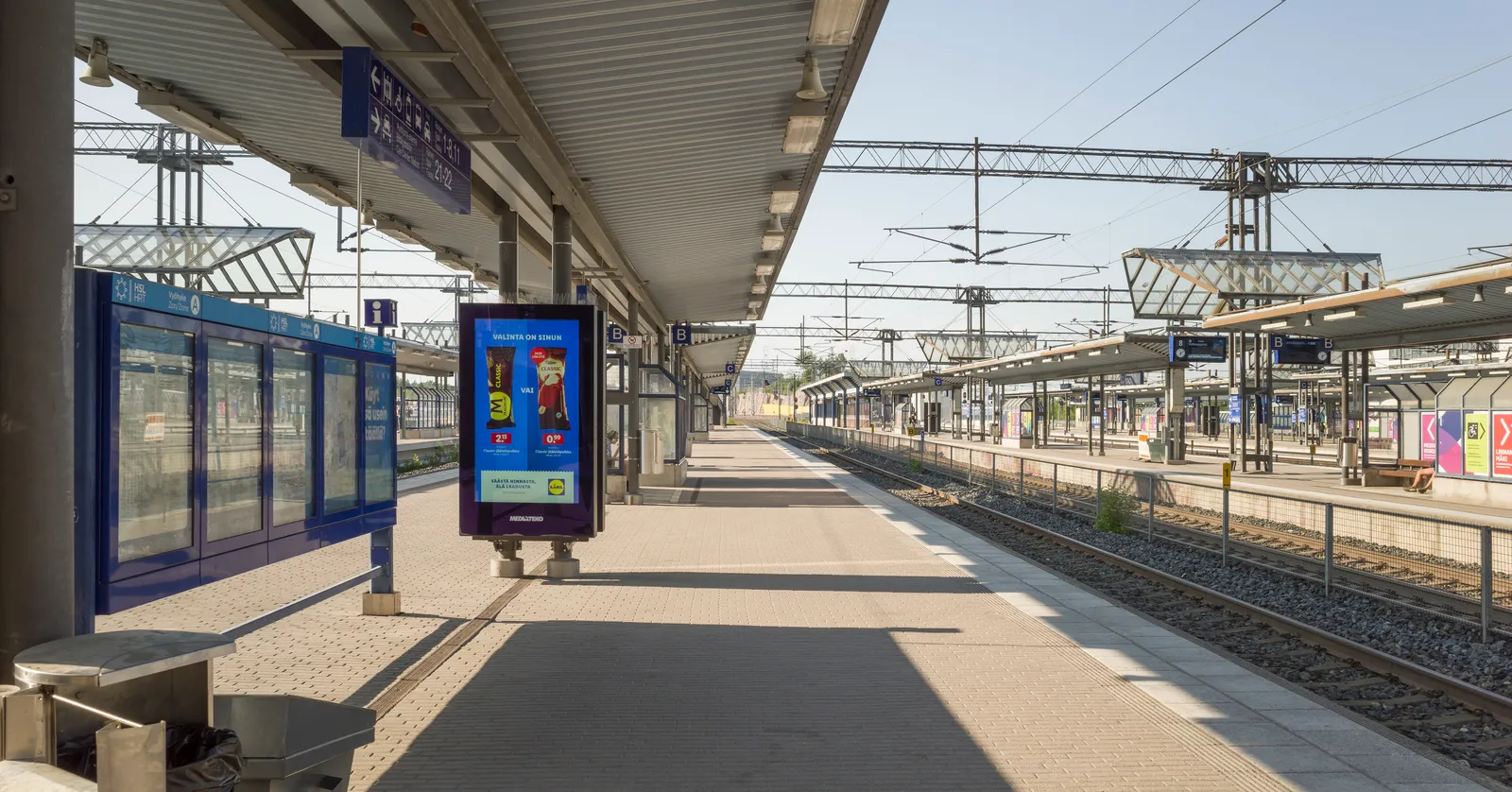 A platform at Pasila station in Helsinki on a clear morning, a long canopy on slim columns over pale paving, blue destination boards hanging from it, and overhead wires running away over several sets of track