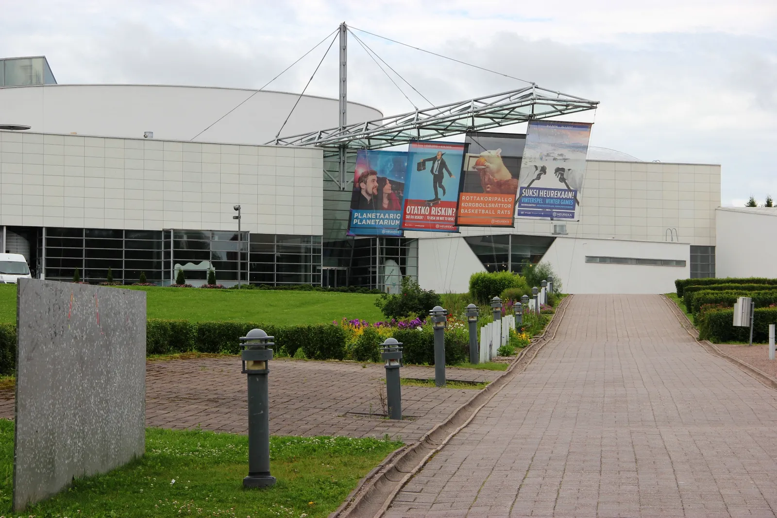 The front of the Heureka science centre in Vantaa, a long low white building with a shallow planetarium dome rising behind it, tall exhibition banners hanging from a steel frame over the entrance and a paved path curving up to the doors
