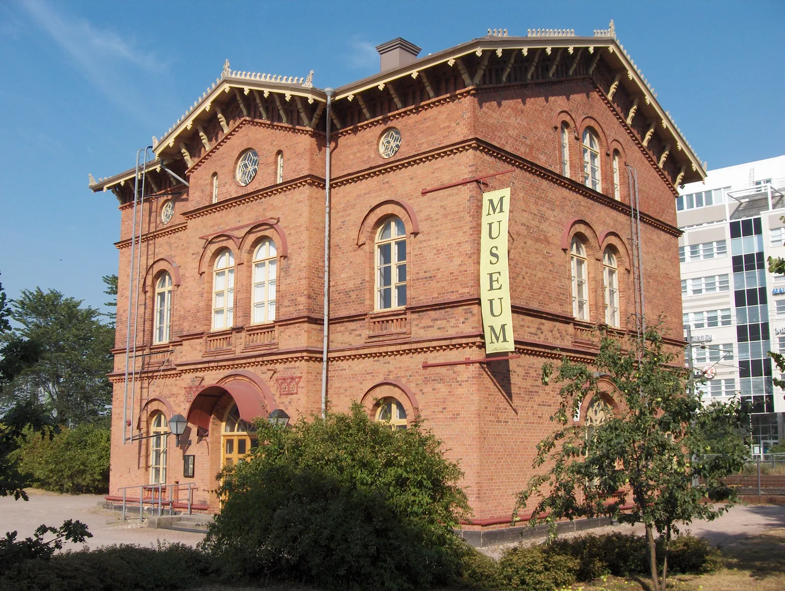 The old Tikkurila station house in Vantaa, a two-storey red brick building with round-arched windows, decorative brick eaves and a small porch, with a yellow banner reading Museum hanging down one corner