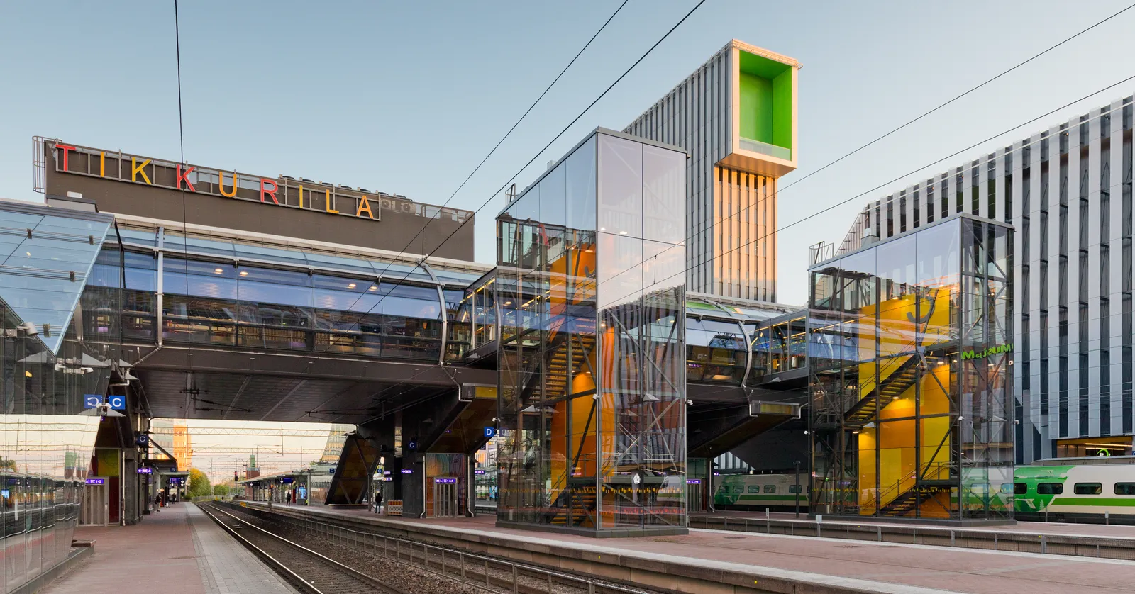Tikkurila station in Vantaa at dusk, the platforms running under a wide overbridge with the station name spelled out in coloured letters along its side, glass lift towers lit orange beside it and a green commuter train standing at the far platform