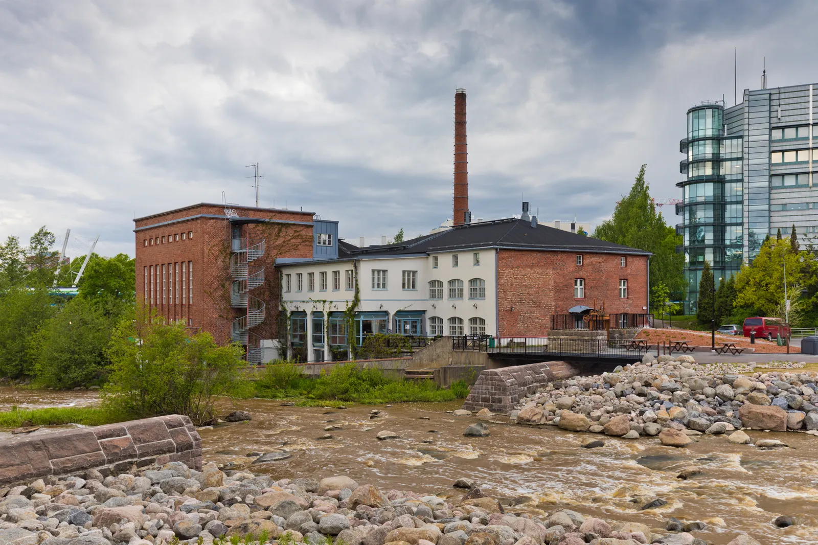 The Vernissa culture factory in Tikkurila, a red brick industrial building with a tall round chimney standing on the bank of the Kerava river, with a bed of pale boulders and shallow water in the foreground