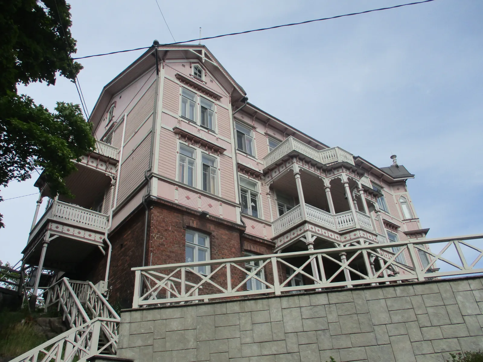 A wooden villa at Linnunlaulu in Helsinki seen from below, three storeys of pale pink timber with white fretwork verandas and balconies on two floors, standing on a brick and granite base above a white painted fence