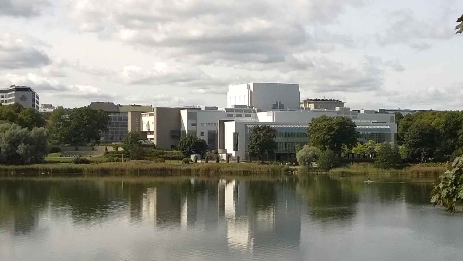 The Finnish National Opera house seen across Töölönlahti bay in Helsinki, a white angular block with lower glazed wings behind a line of trees, the whole building reflected in still water