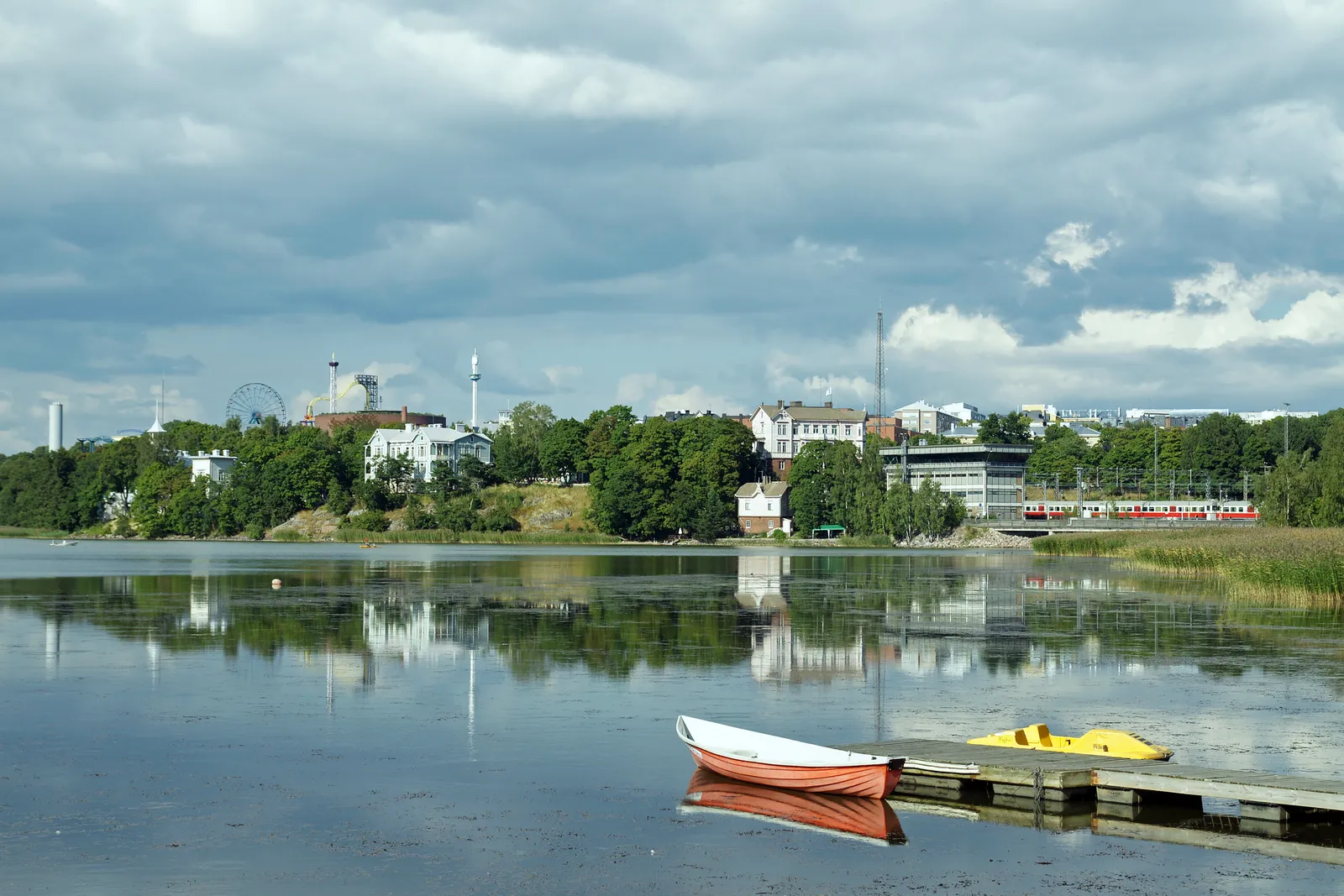 Töölönlahti bay in Helsinki from the water's edge, a white rowing boat and a yellow pedal boat tied to a low jetty in the foreground, reeds along the near shore and a wooded bank with pale buildings on the far side