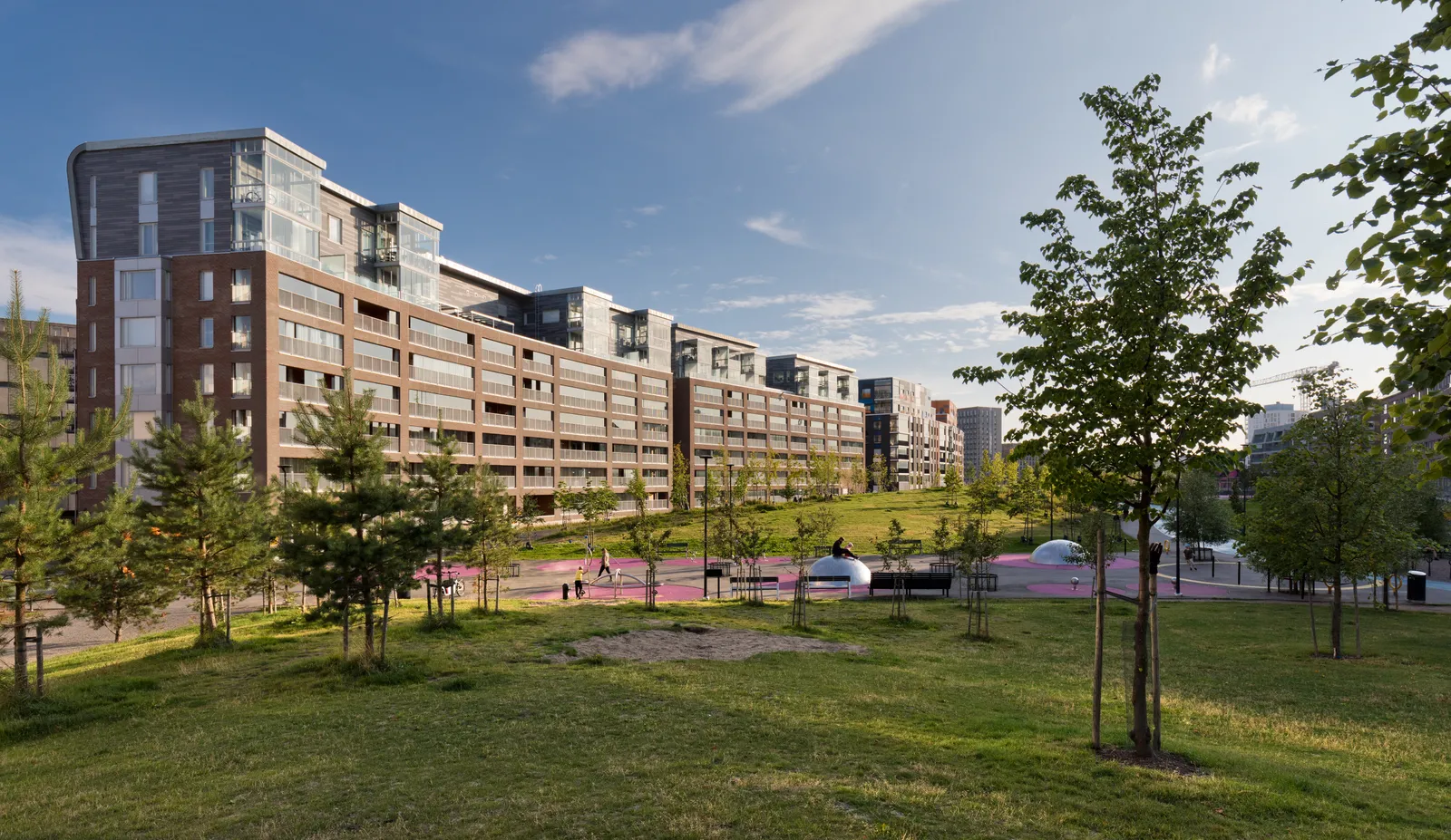 Hyväntoivonpuisto park in Jätkäsaari, Helsinki, young trees and mown grass in the foreground with a red-surfaced walking and cycling path curving through it and a long row of new brick and glass apartment blocks along one side