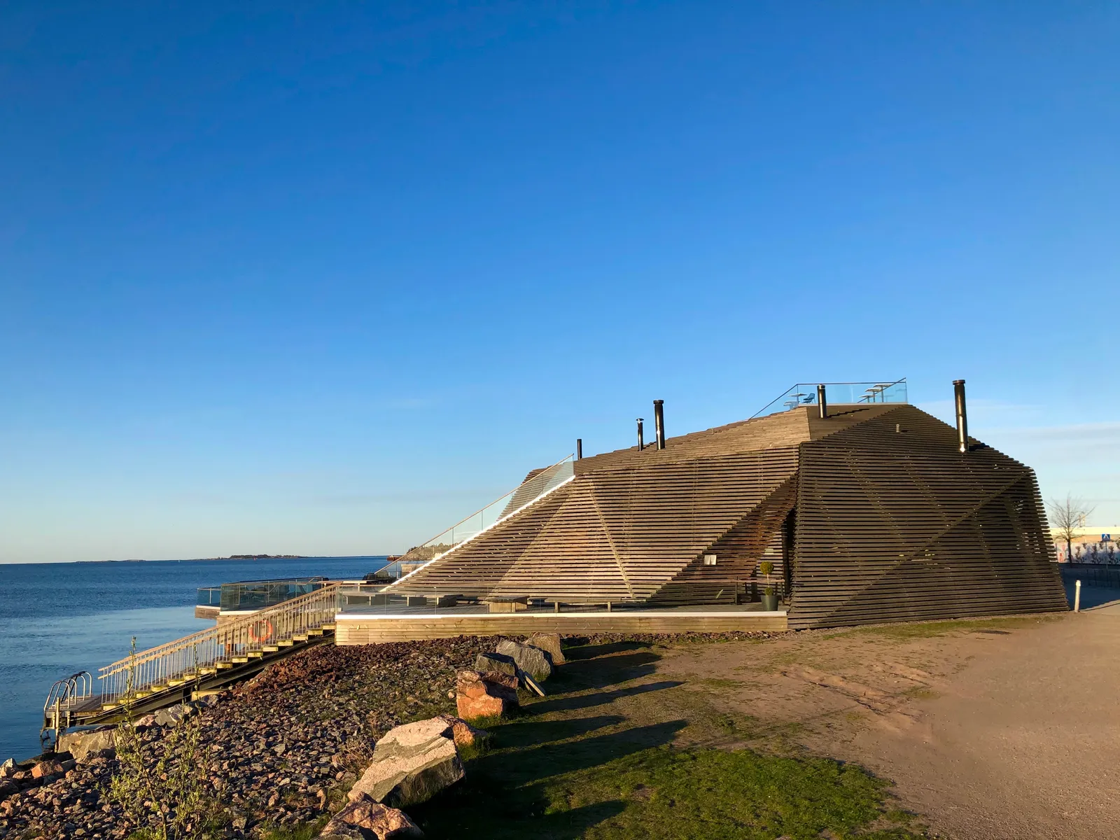 The Löyly sauna building on the Helsinki shoreline, a low mound clad in angled timber slats with a public staircase running up its side to a roof terrace, standing on a rocky edge beside open sea