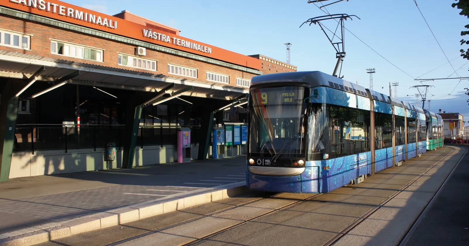 A blue and white tram waiting at the West Terminal stop in Helsinki, with the long brick terminal building behind it carrying the words Länsiterminaali and Västra terminalen above a glazed canopy