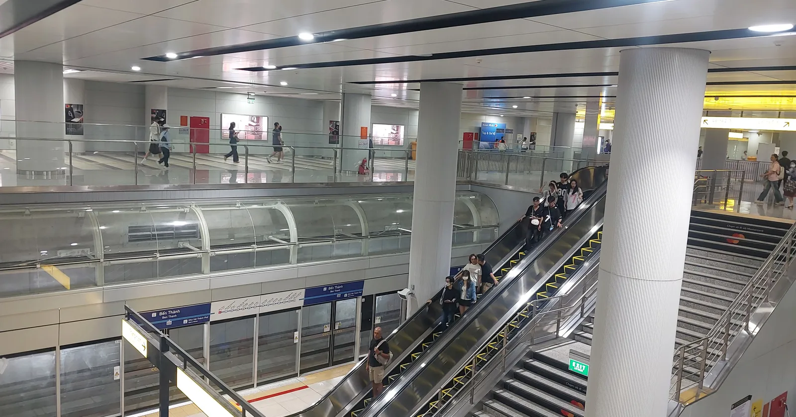 The underground concourse of Ben Thanh station in Ho Chi Minh City, banks of escalators running down between white columns to the platform level with passengers on the walkway above
