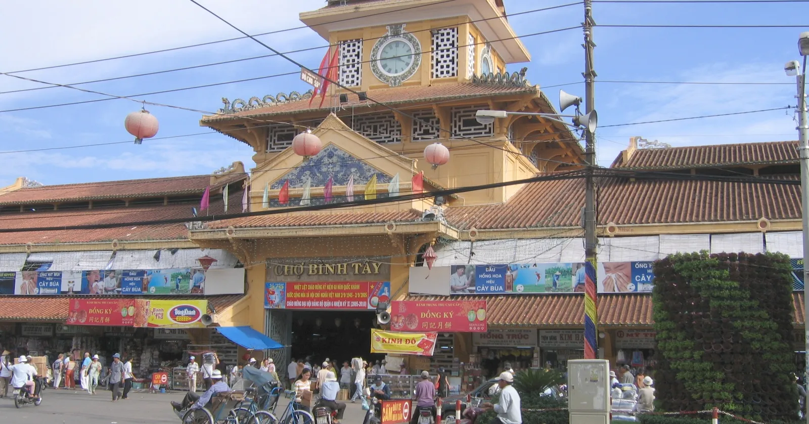 The main gate of Binh Tay Market in Cho Lon, a tiered yellow tower with a clock face under a curved tiled roof, red lanterns strung across the front and shoppers crowding the entrance below