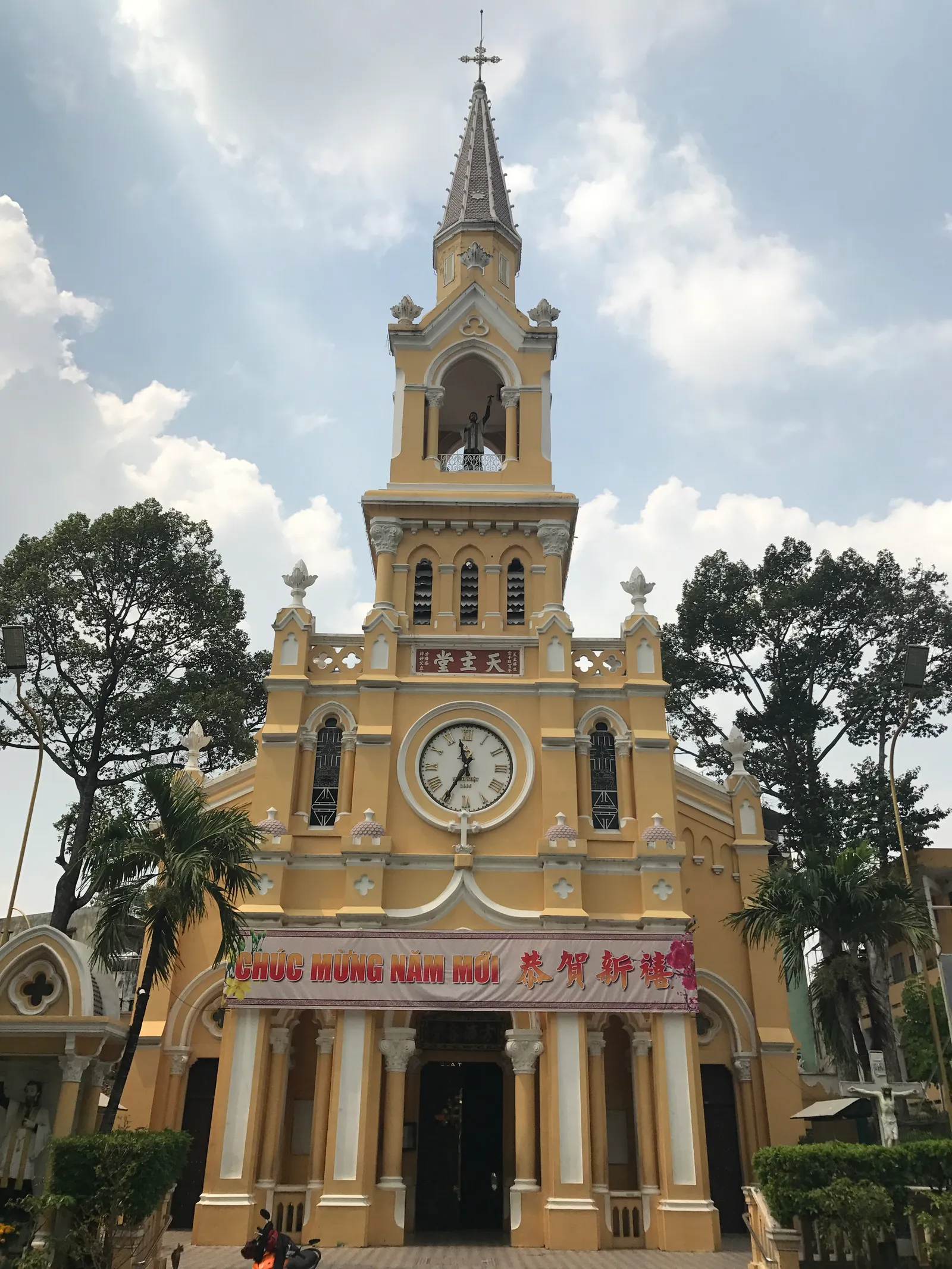 The yellow frontage of Cha Tam Church in Cho Lon, a clock set into the gable below a slim spire, with Chinese characters on a red panel above the main door