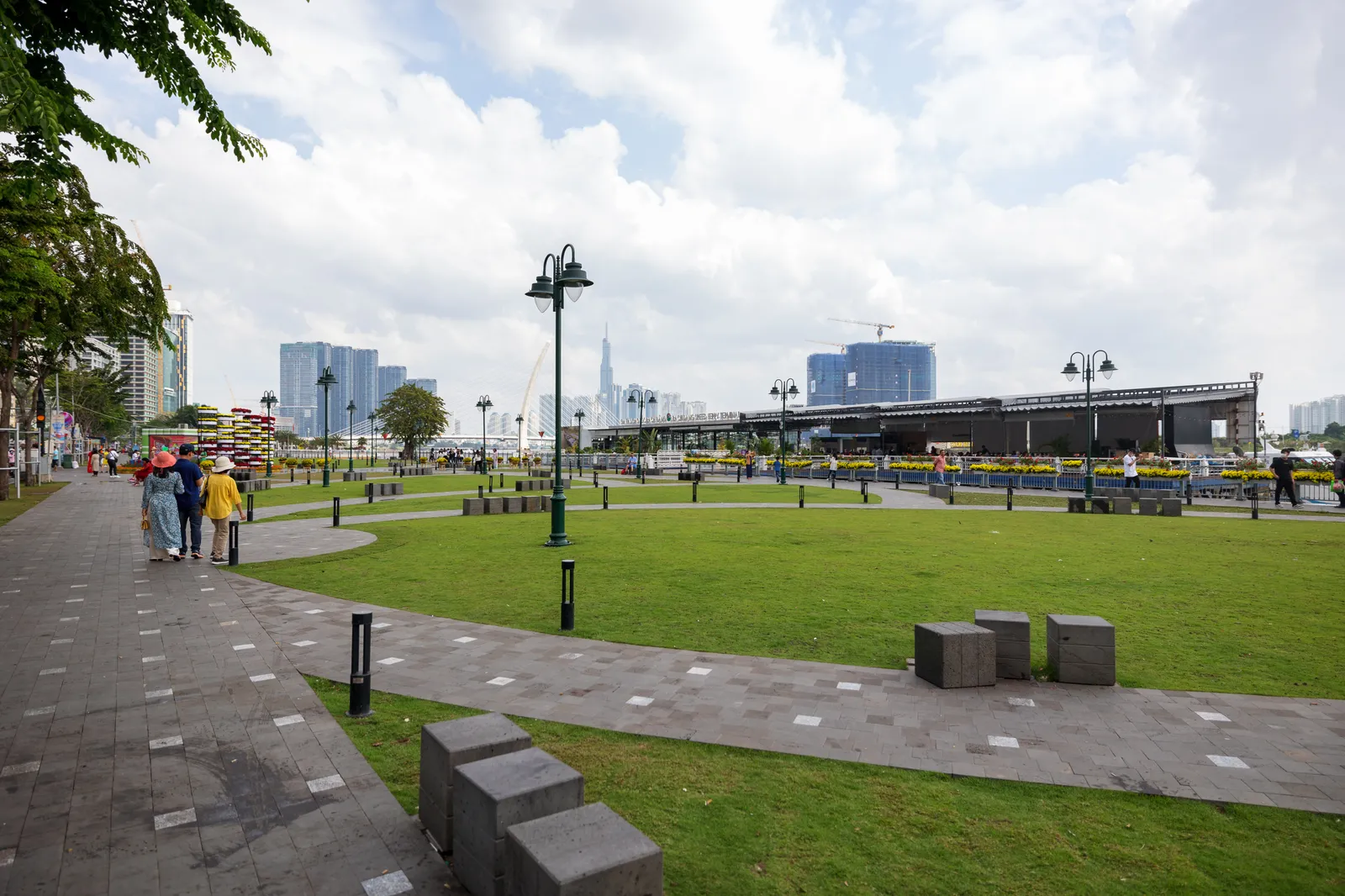 The lawns and paved paths of Bach Dang wharf park along the Saigon River, with lamp posts, low stone blocks and the city skyline behind the trees