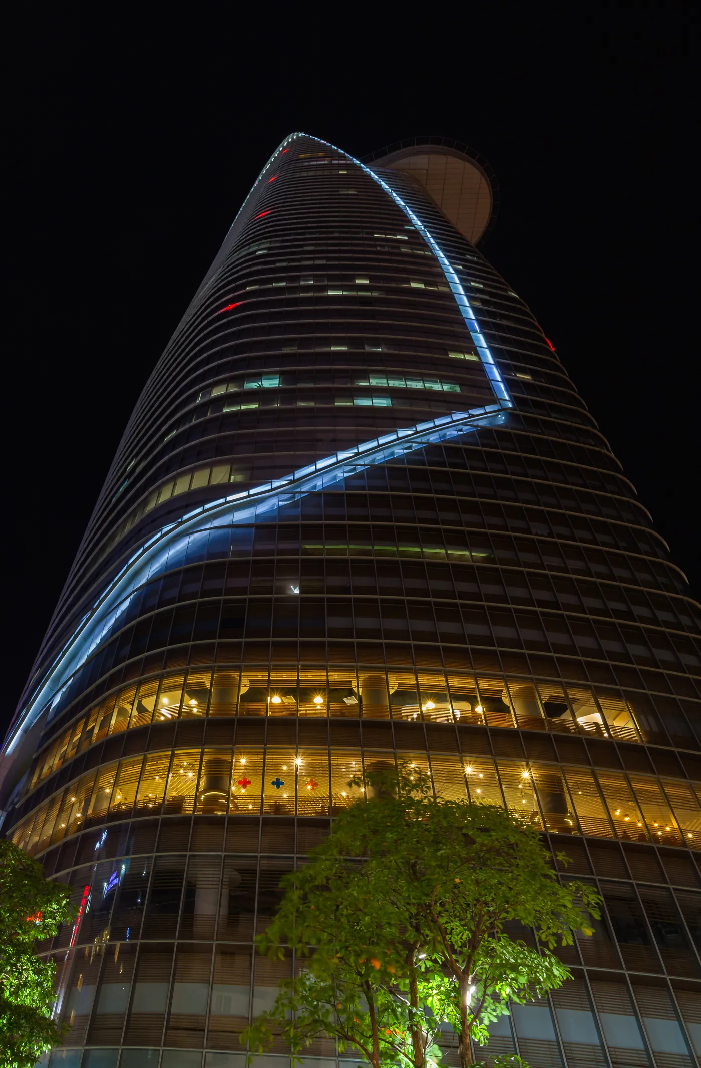Bitexco Financial Tower in Ho Chi Minh City photographed from its base at night, the curved glass flank lit blue and the helipad jutting out from the side near the top