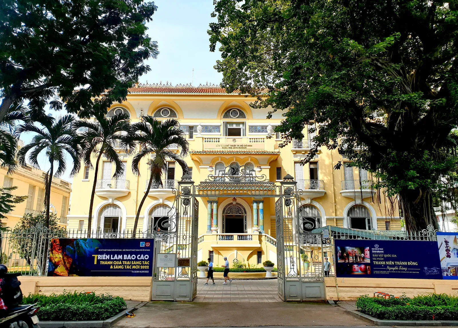 The yellow mansion housing the Ho Chi Minh City Museum of Fine Arts, seen through its white wrought-iron gates with palms in the forecourt and exhibition banners along the railings
