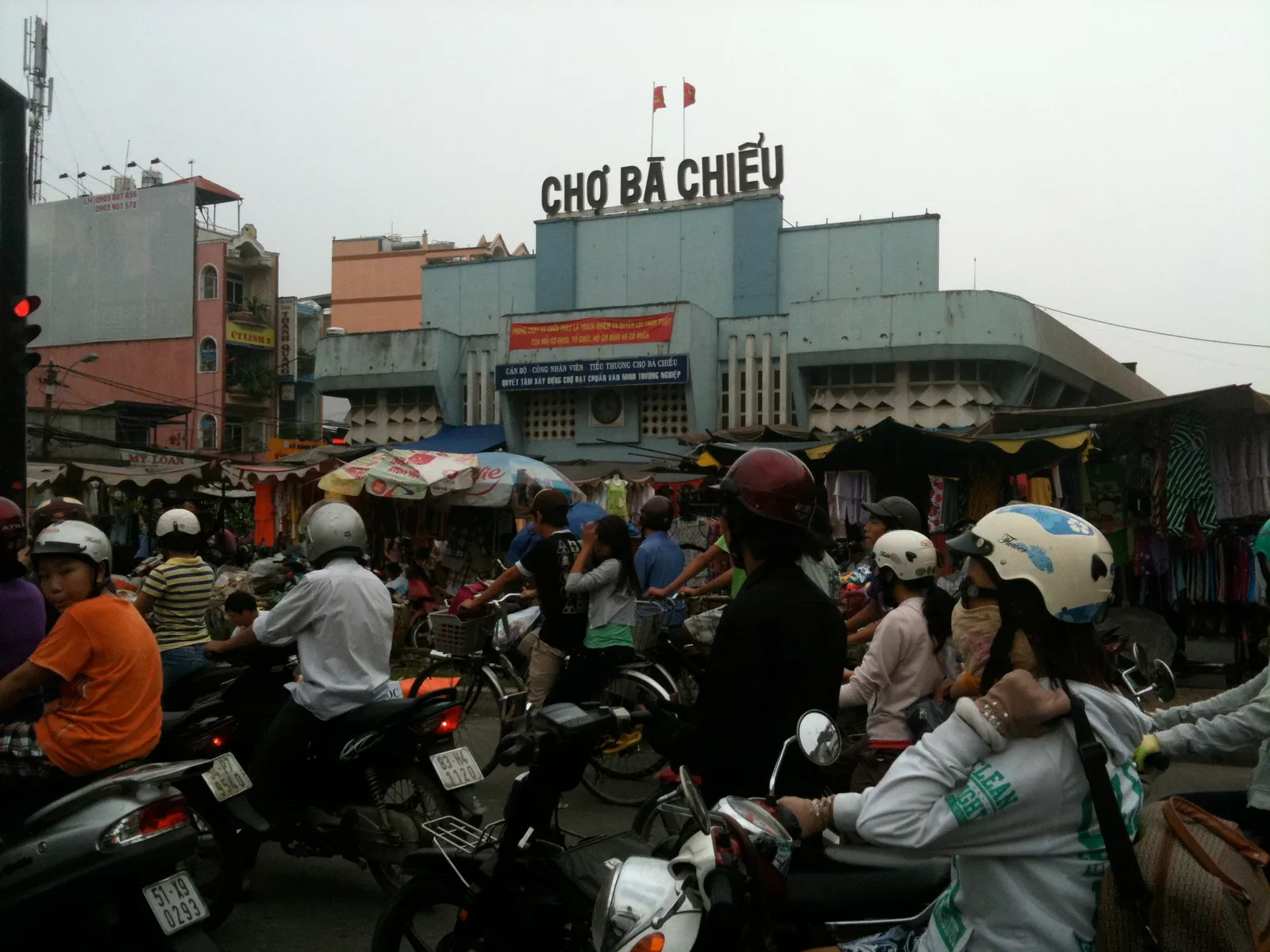 The frontage of Cho Ba Chieu market in Binh Thanh with its name in tall red letters along the roofline, stalls under umbrellas below and motorbikes filling the junction in front