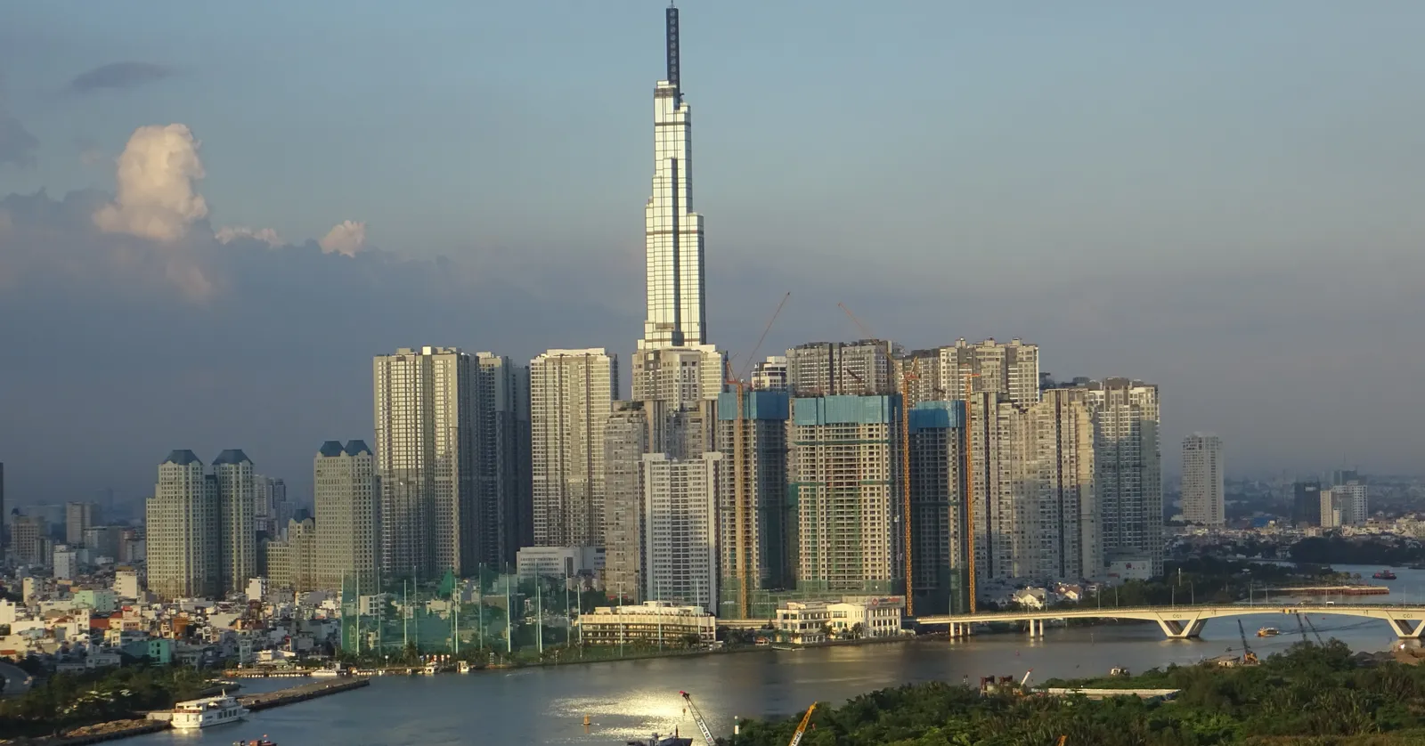Landmark 81 rising in a spire above the cluster of Vinhomes Central Park apartment towers on the Saigon River in Binh Thanh, with the river and a bridge in the foreground