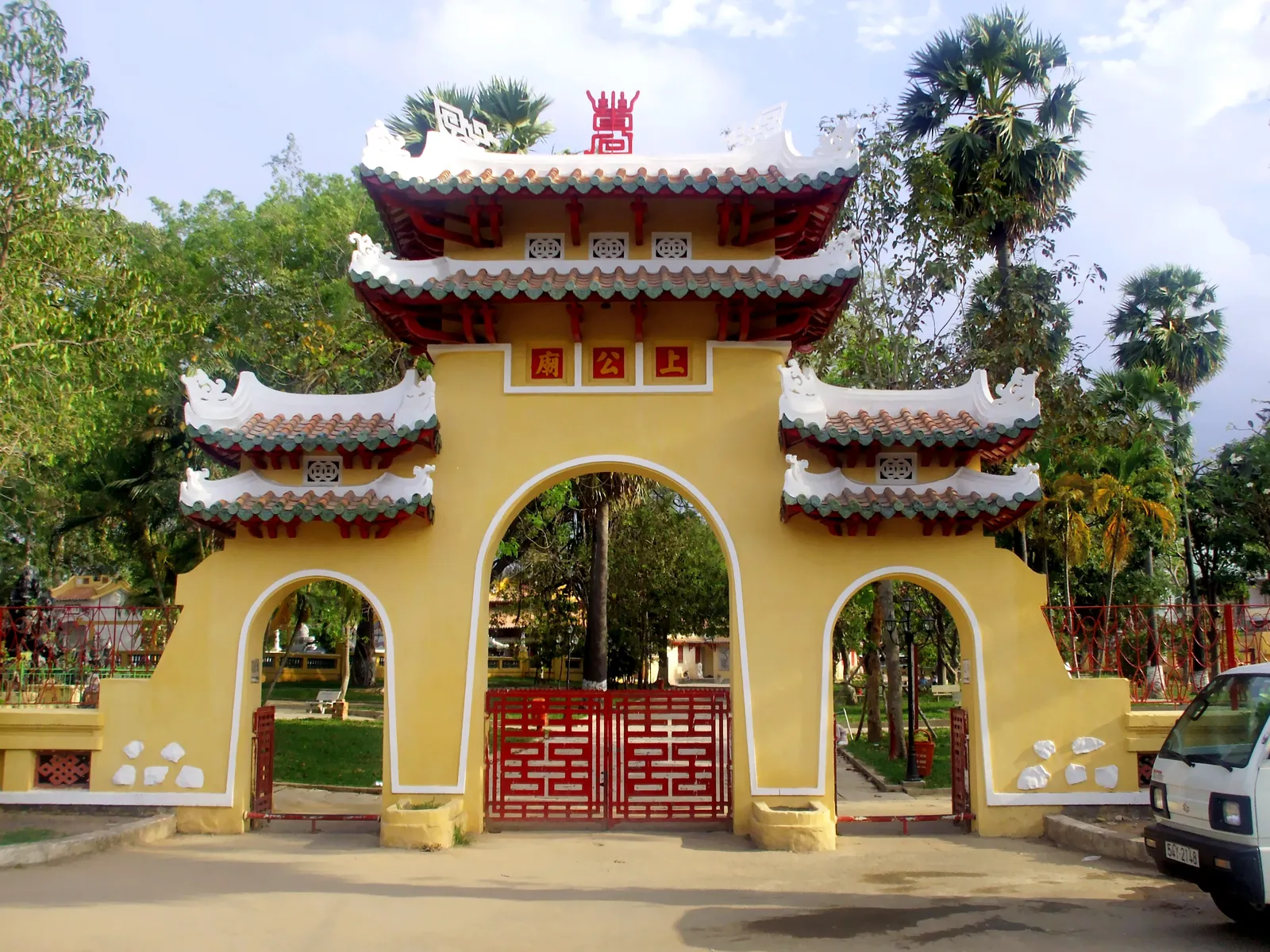 The yellow triple-arched gate of Le Van Duyet's tomb in Ba Chieu, its tiled roofs stacked in three tiers above red lattice doors, with palms and trees in the grounds behind