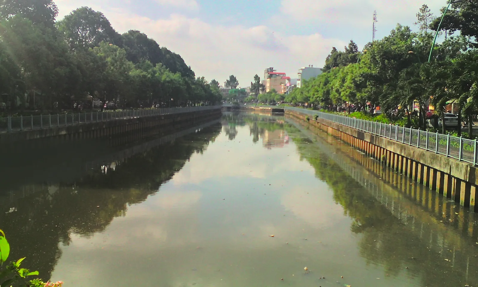 The Nhieu Loc-Thi Nghe canal in Ho Chi Minh City seen from a bridge, still water between two railed embankments with lines of mature trees along both banks