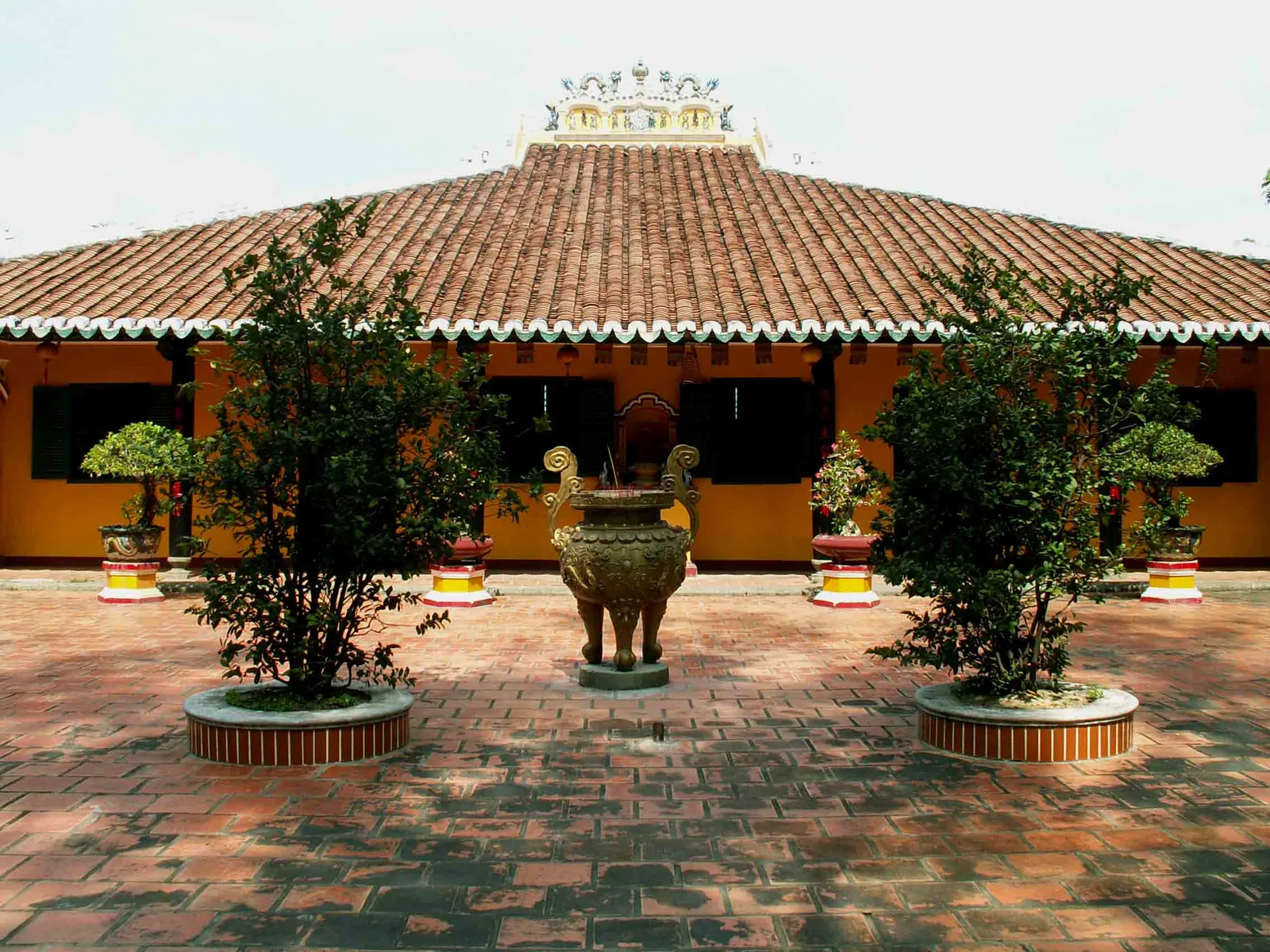 The tiled roof and brick courtyard of Giac Lam Pagoda in Tan Binh, a large bronze incense urn standing between two clipped trees in front of the ochre timber hall