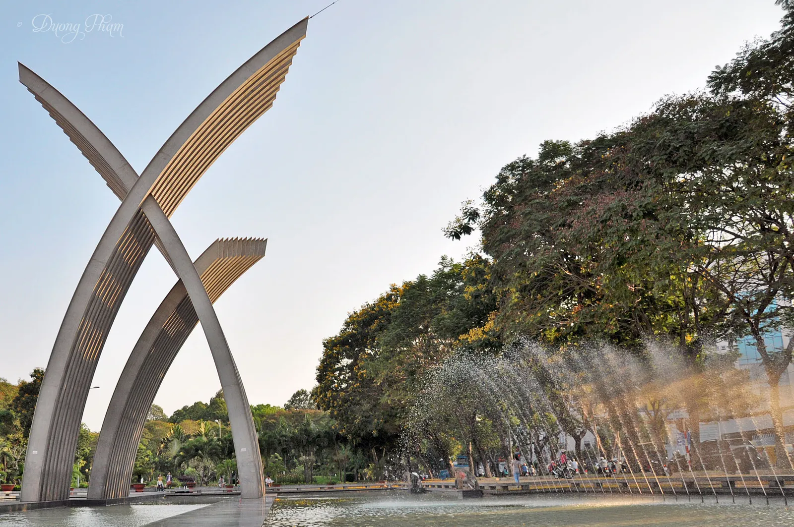 The crossed steel arches at Hoang Van Thu Park in Tan Binh rising beside the park's lake, with a fountain jetting across the water and mature trees along the far bank