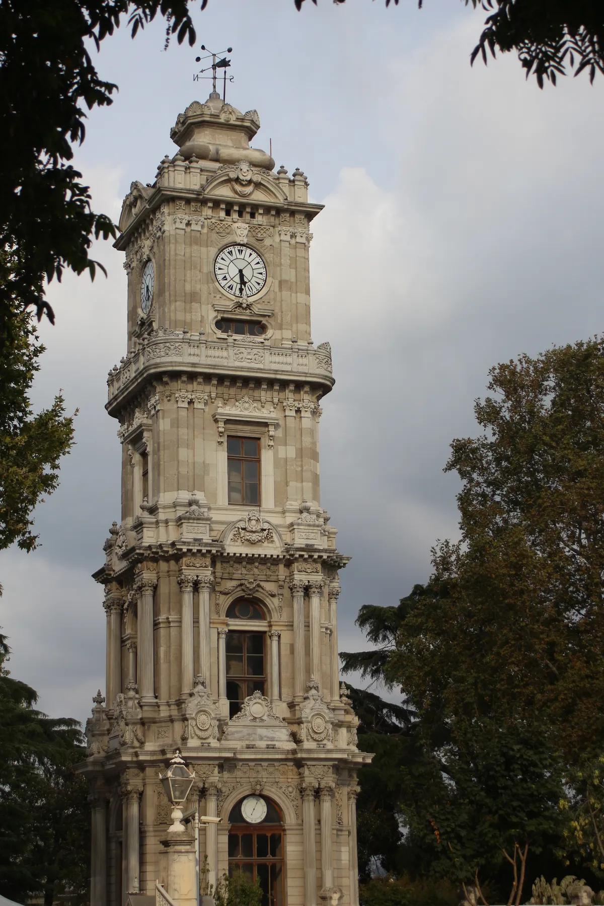 The Dolmabahçe Clock Tower in Istanbul, a four-storey stone tower in ornate 19th-century style with a white clock face near the top, framed by trees against a grey sky