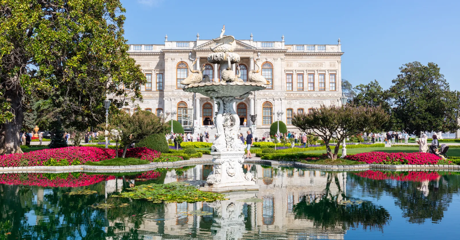 The garden front of Dolmabahçe Palace in Istanbul, a long white neoclassical facade with carved pediments and tall windows, behind a formal pool with a marble fountain and beds of pink flowers
