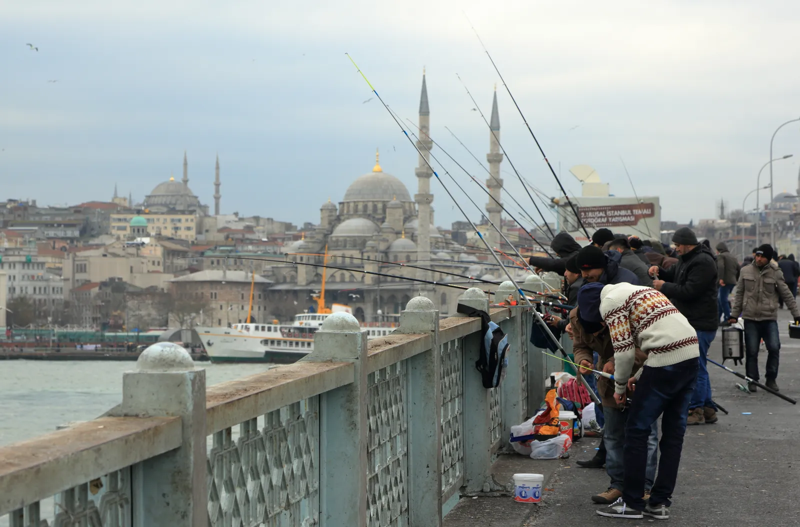 A row of men fishing over the rail of the Galata Bridge in Istanbul, rods angled out over the water, with ferries below and the domes and minarets of the New Mosque across the Golden Horn behind them