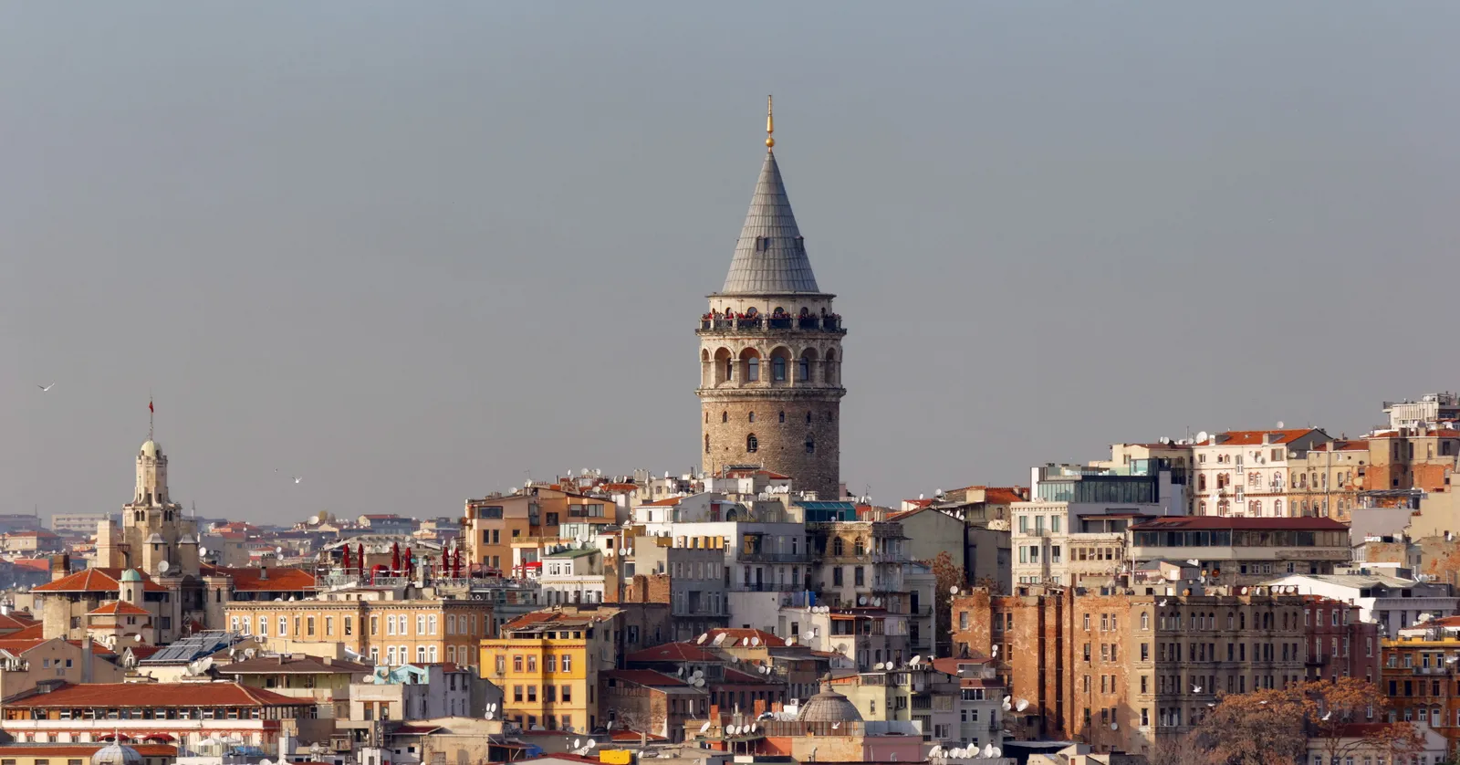 The Galata Tower rising above the rooftops of Istanbul, a round stone drum with a conical grey roof and a railed balcony below it, surrounded by close-packed apartment blocks running down the hill