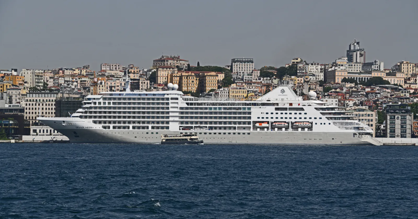 A white cruise ship tied up along the Galataport waterfront in Istanbul, seen from the Bosphorus, with the hillside houses of Beyoğlu rising behind