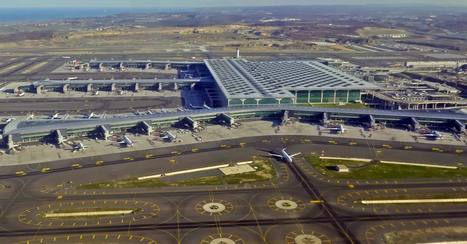 Istanbul Airport from the air, the long dark roof of the single terminal building stretching across the frame with aircraft parked at piers on both sides and taxiways marked out in yellow across the foreground