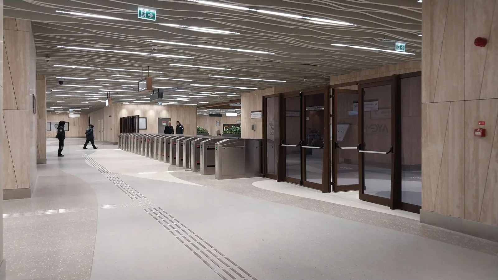 The metro station under Istanbul Airport, a wide stone-clad concourse with a row of ticket gates along one side and glass doors leading through to the platforms