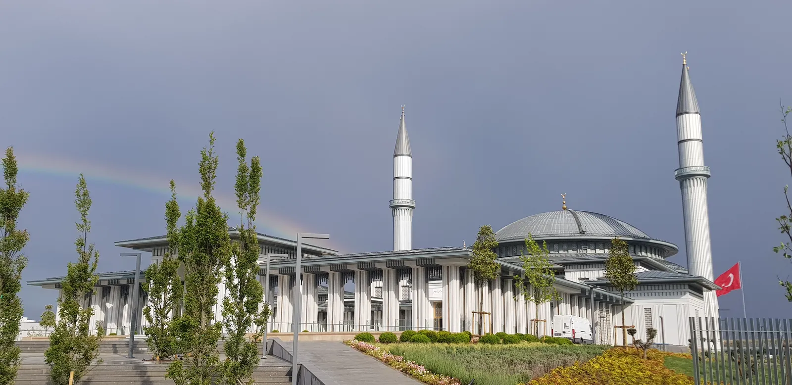 Ali Kuşçu Mosque outside Istanbul Airport, a low white colonnaded building with a wide shallow dome and two slim minarets, with young trees and flowerbeds in front of it under a grey sky