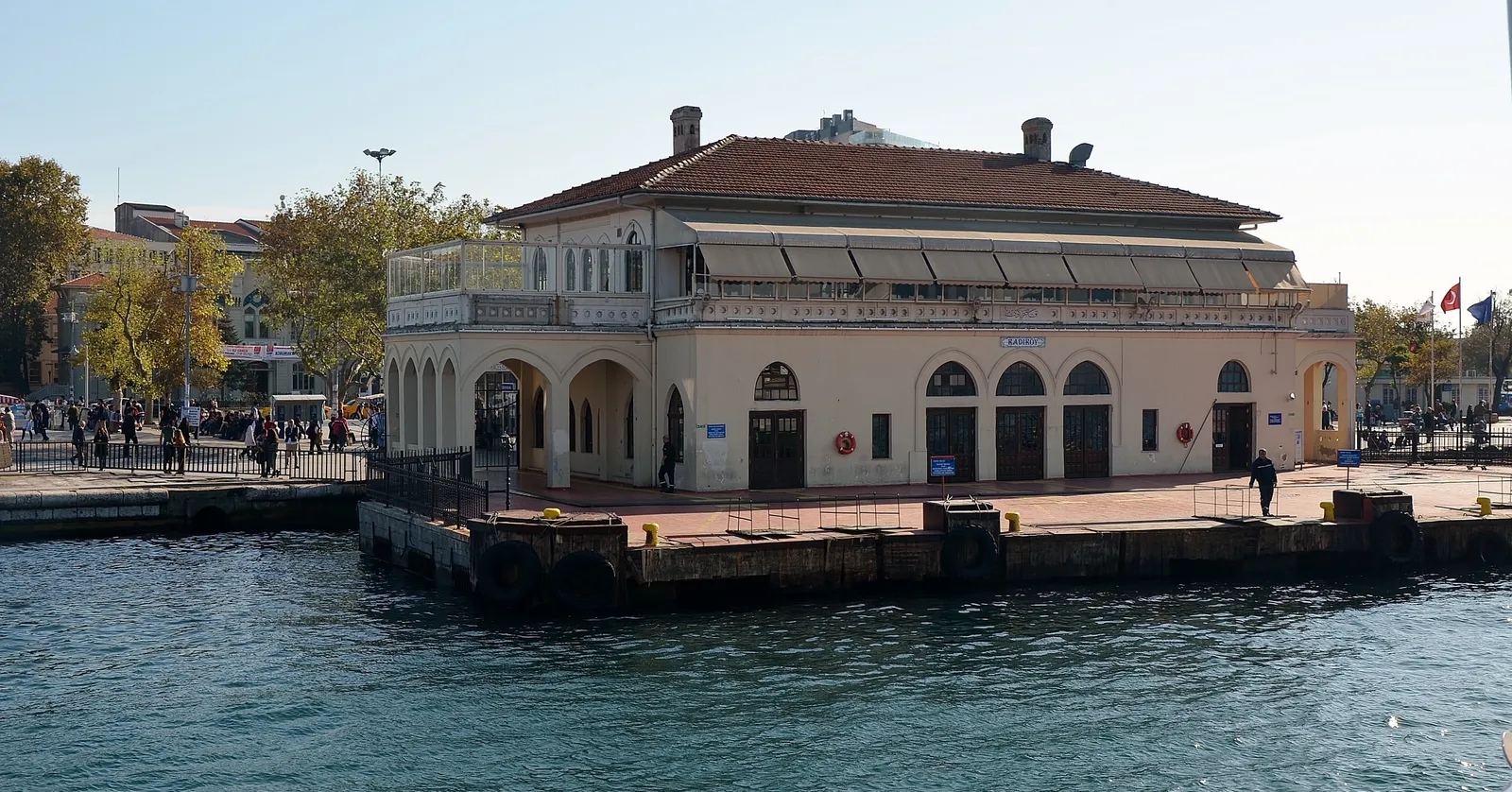 The old Kadıköy ferry pier in Istanbul seen from the water, a low stone terminal building with arched openings and a tiled roof standing out on its own jetty, with people on the waterfront behind it
